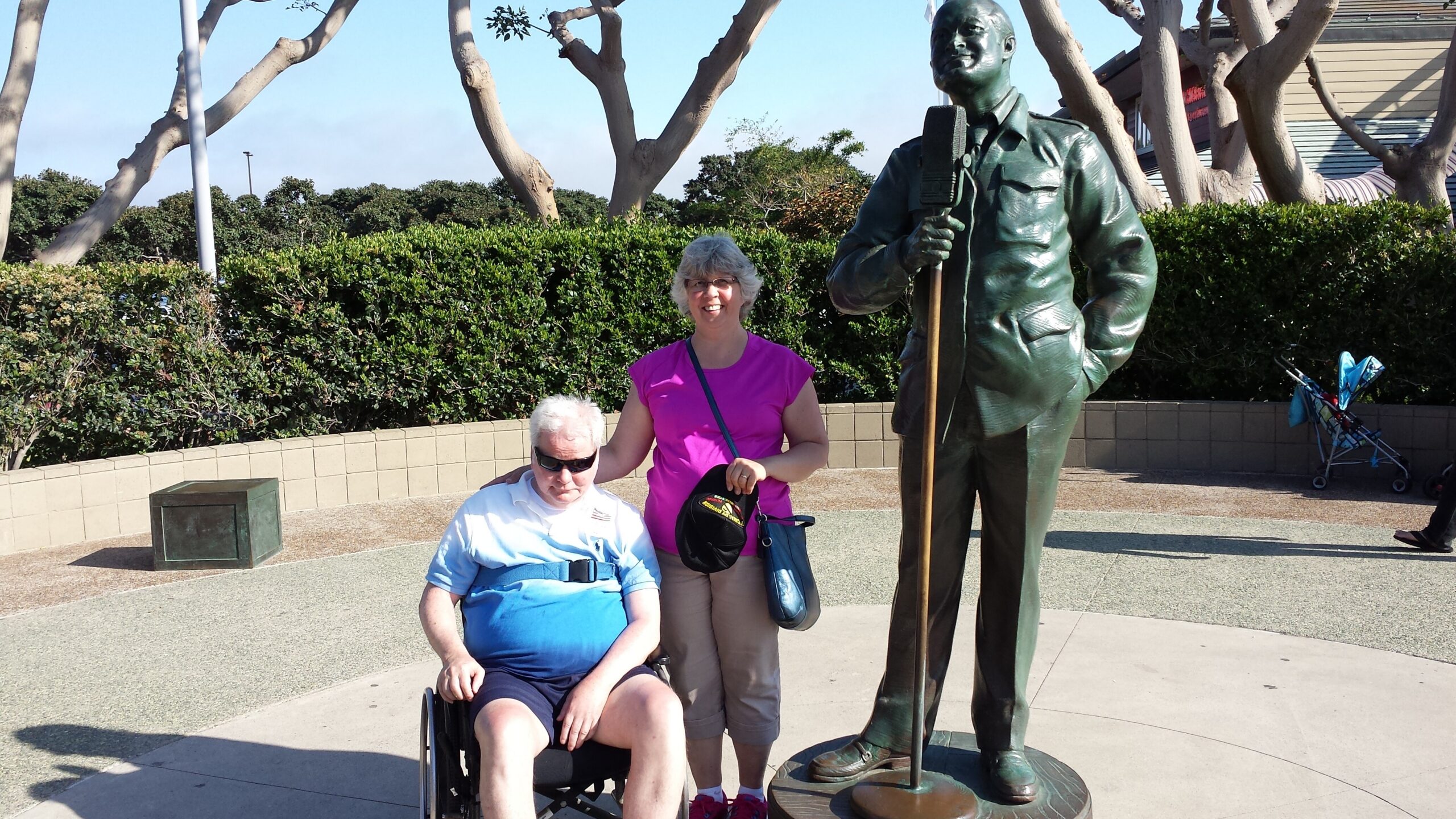 Woman in pink shirt stands beside an elderly man in a wheelchair, posing next to a tall bronze statue holding a microphone outdoors with shrubbery in the background.
