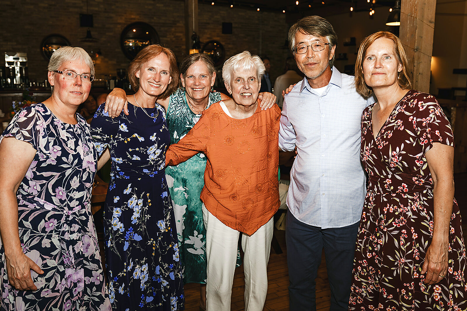 Six adults posing together for a photo at a warmly lit social event, wearing colorful floral and patterned dresses and a light check shirt. They smile at the camera in a casual group shot.
