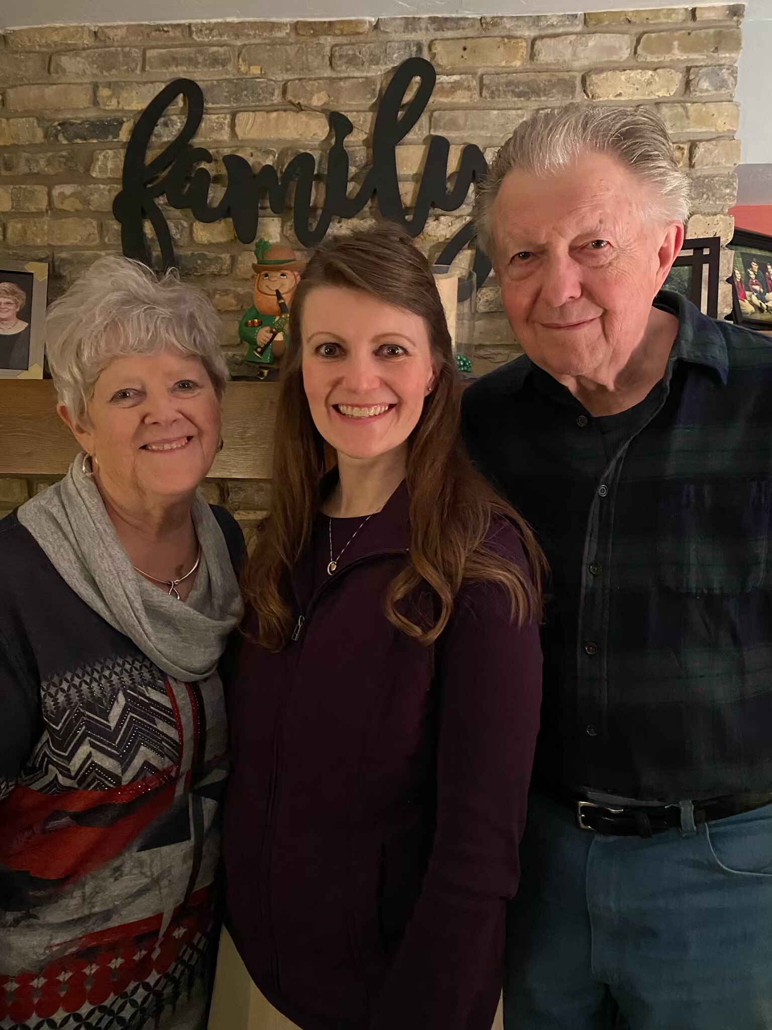 Three people pose for a warm group photo indoors, standing in front of a brick wall with a decorative 'family' sign.
