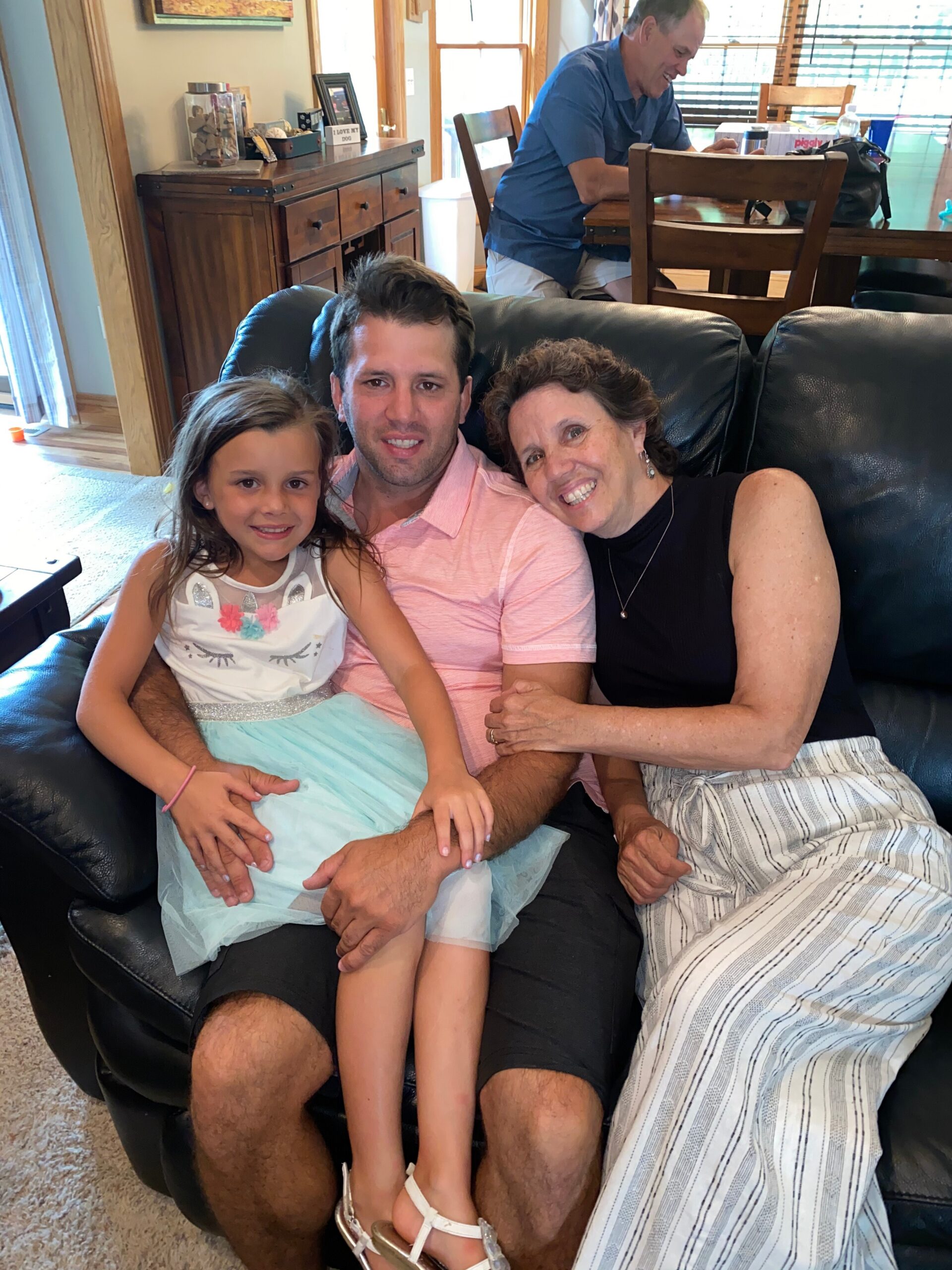 Family of three on a sofa: dad, daughter, and grandma smiling together; a man sits at a table in the background.