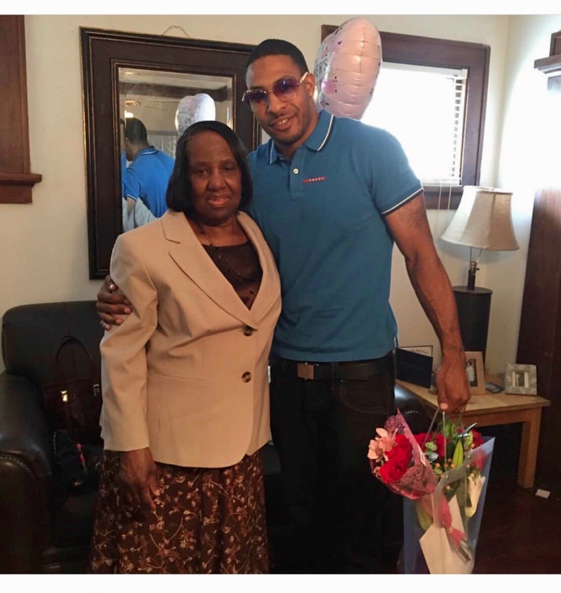 Man wearing sunglasses and a teal polo stands beside an elderly woman in a beige blazer inside a living room, holding a bouquet of flowers and with a pink balloon nearby.