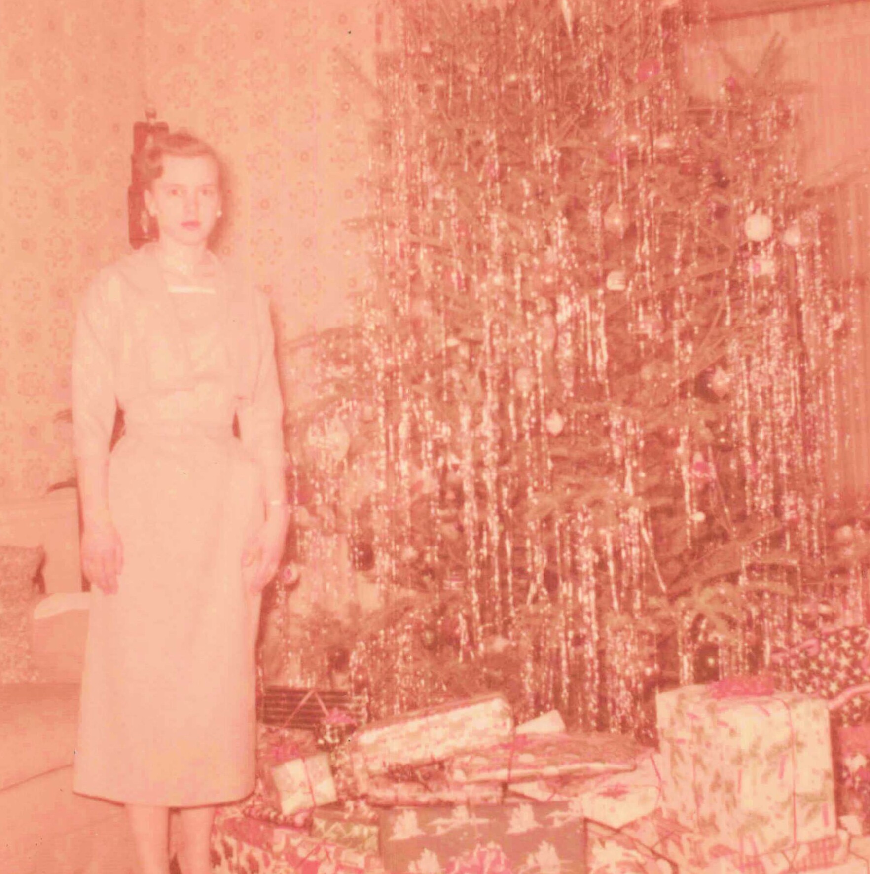 Woman in a pale dress standing indoors beside a large decorated Christmas tree in a vintage photo.