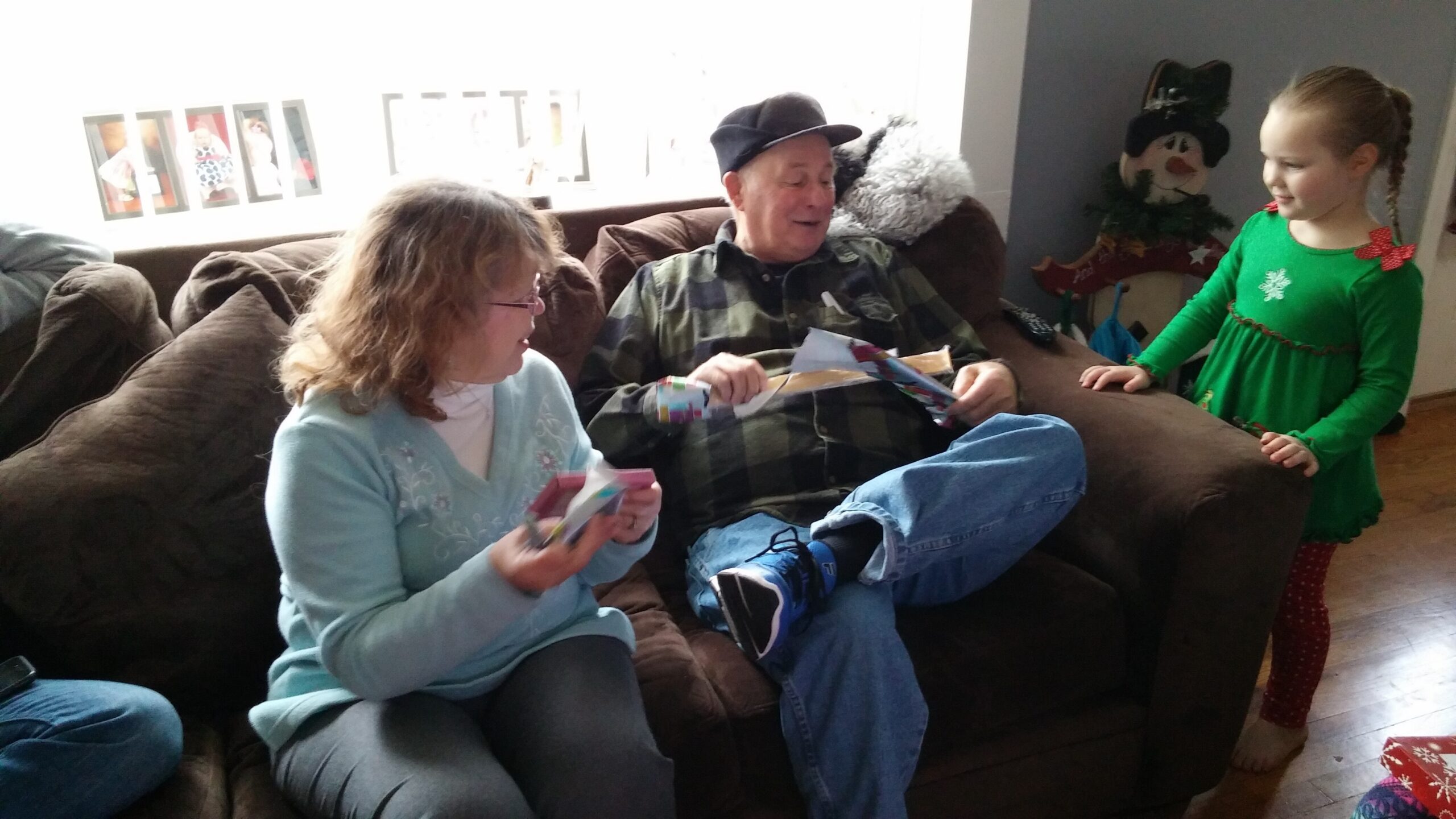 Older man in a plaid shirt sits on a brown couch opening gifts as two women hand him presents; a girl in a green Christmas dress stands nearby, smiling.