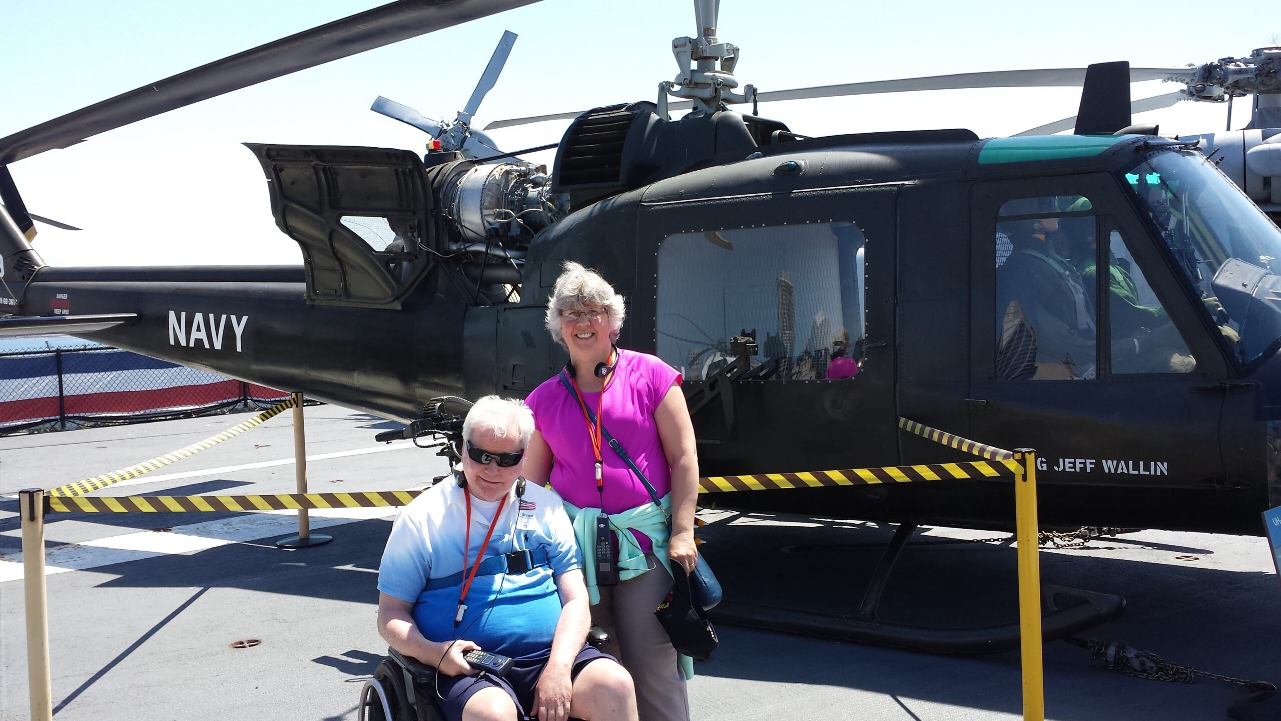 Two smiling visitors pose in front of a dark navy helicopter on display, one in a wheelchair with a vest and the other standing beside it.