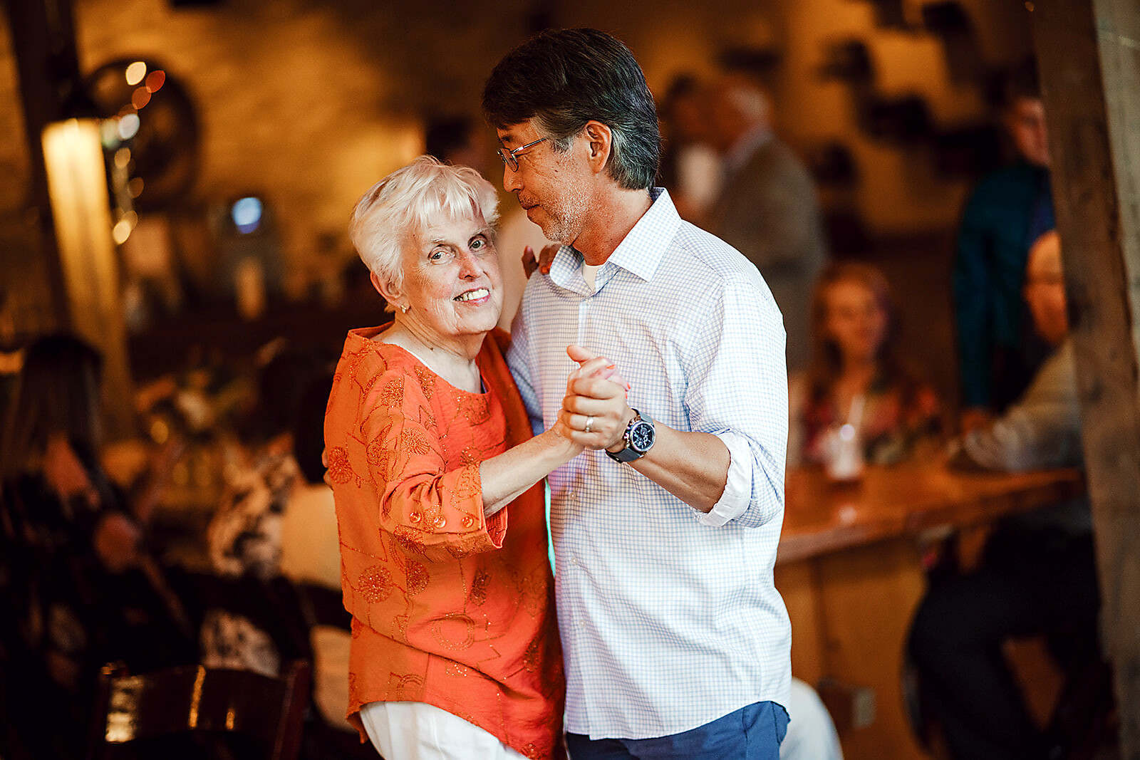 Smiling elderly woman in an orange top dancing with a man in a checked shirt at a warmly lit party.