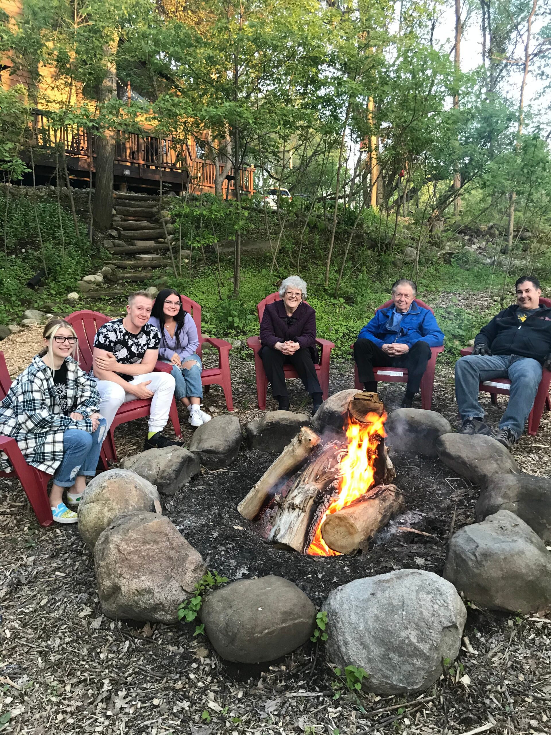 Group of six people seated in red chairs around a backyard campfire ring with rocks in a wooded yard behind them
