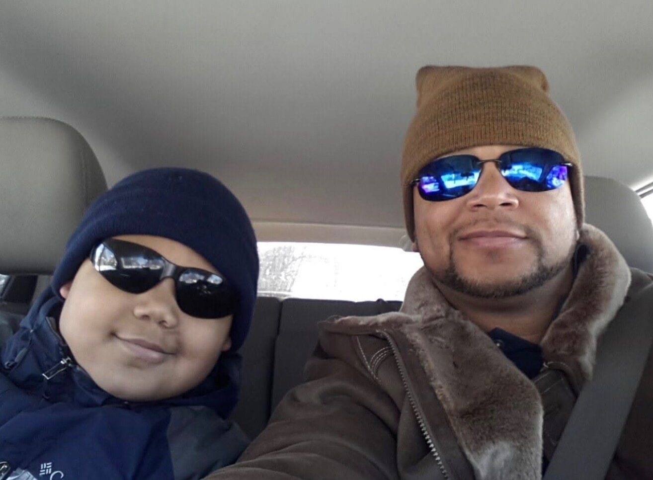 Two people wear sunglasses and warm hats, smiling at the camera inside a car.