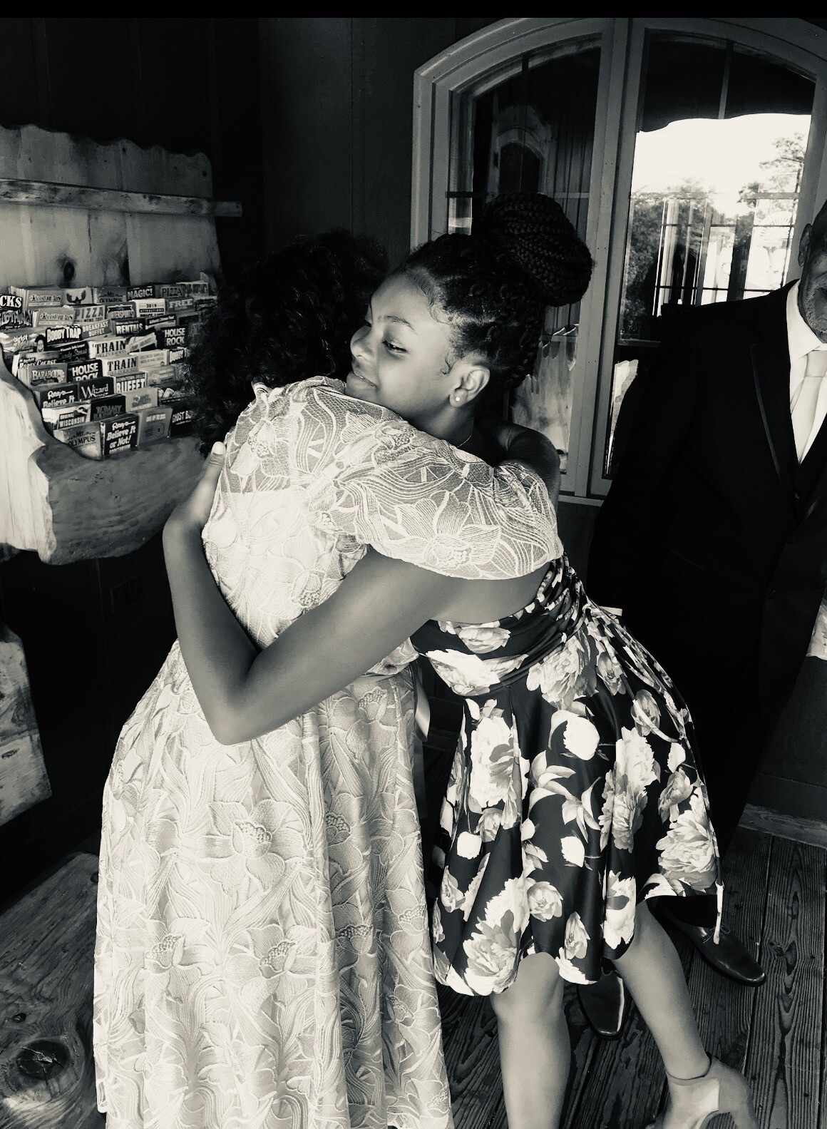 Two people share a warm hug at an event, one in a lace dress and the other in a floral dress, smiles visible in a candid moment indoors.