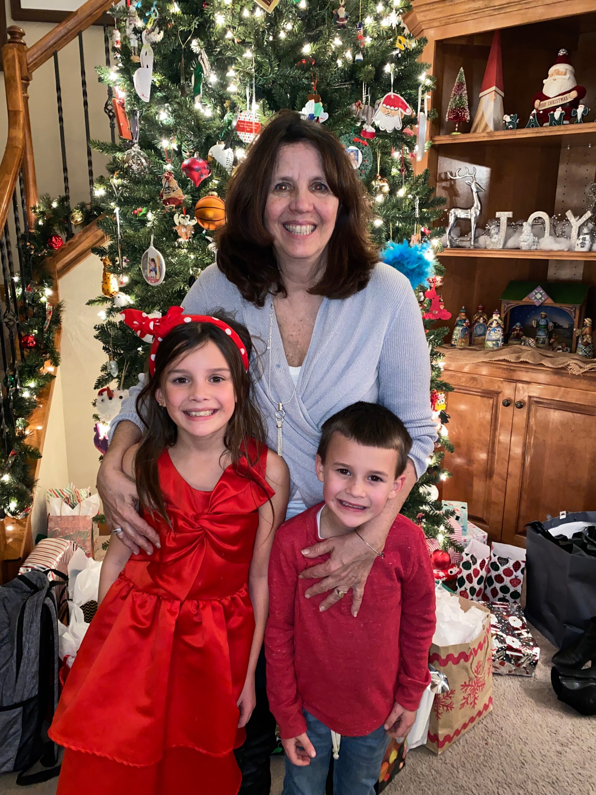 Smiling woman with two children in festive red outfits posing by a lit Christmas tree with gift bags around them in a living room.