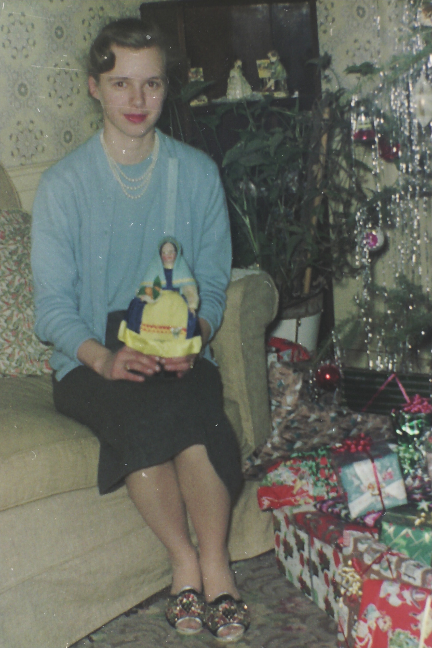Woman in a light blue sweater and pearl necklace sits on a sofa, holding a yellow toy or basket as a Christmas tree and wrapped gifts appear nearby.