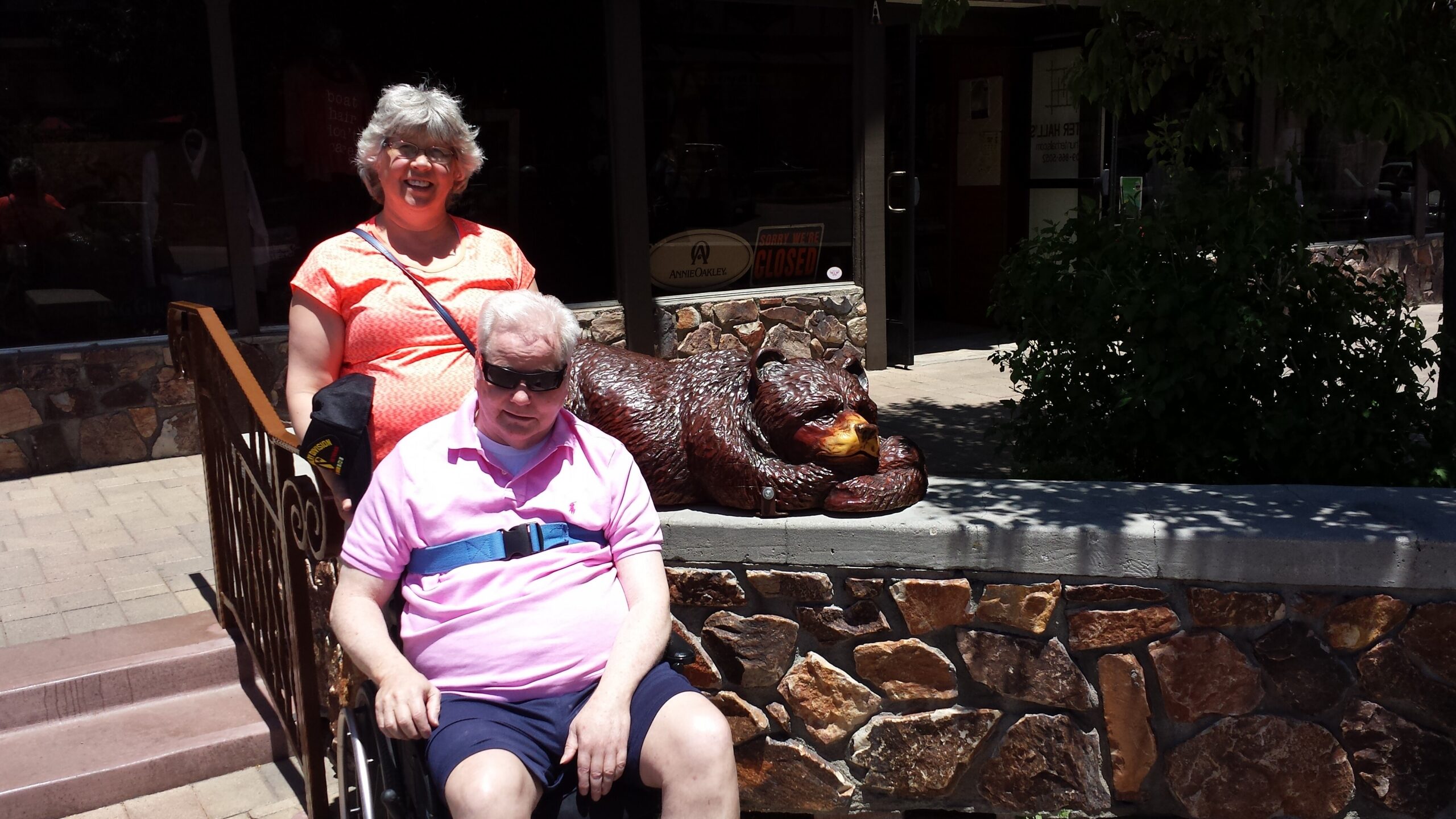 Smiling woman in an orange shirt stands beside a man in a wheelchair with a bear sculpture and storefront in the background.