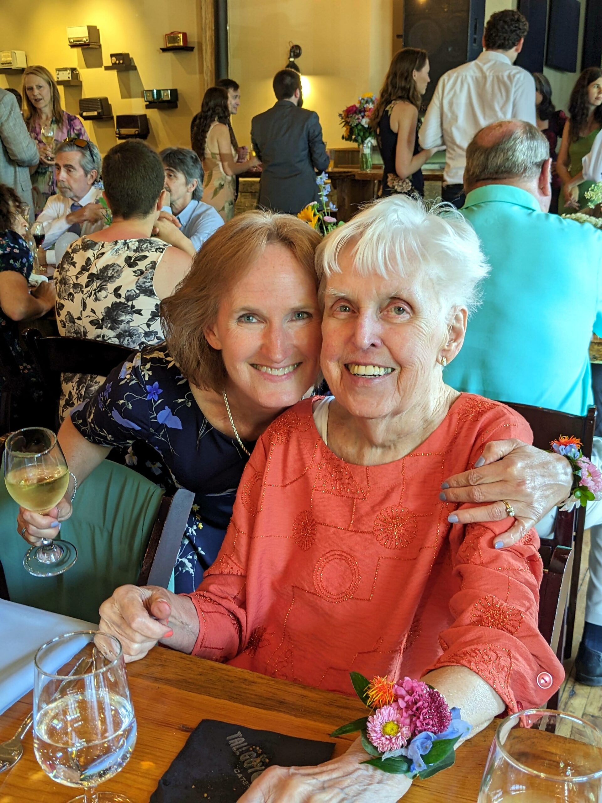 Two smiling women sit close together at a festive dinner table, one in coral with a corsage and the other in a blue floral dress, with flowers and glasses in the foreground.