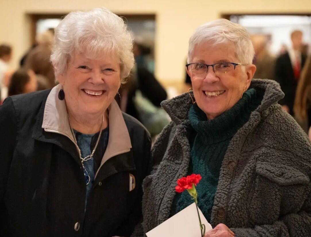 Two smiling elderly women stand together at a social event, one holding a red carnation and a card.