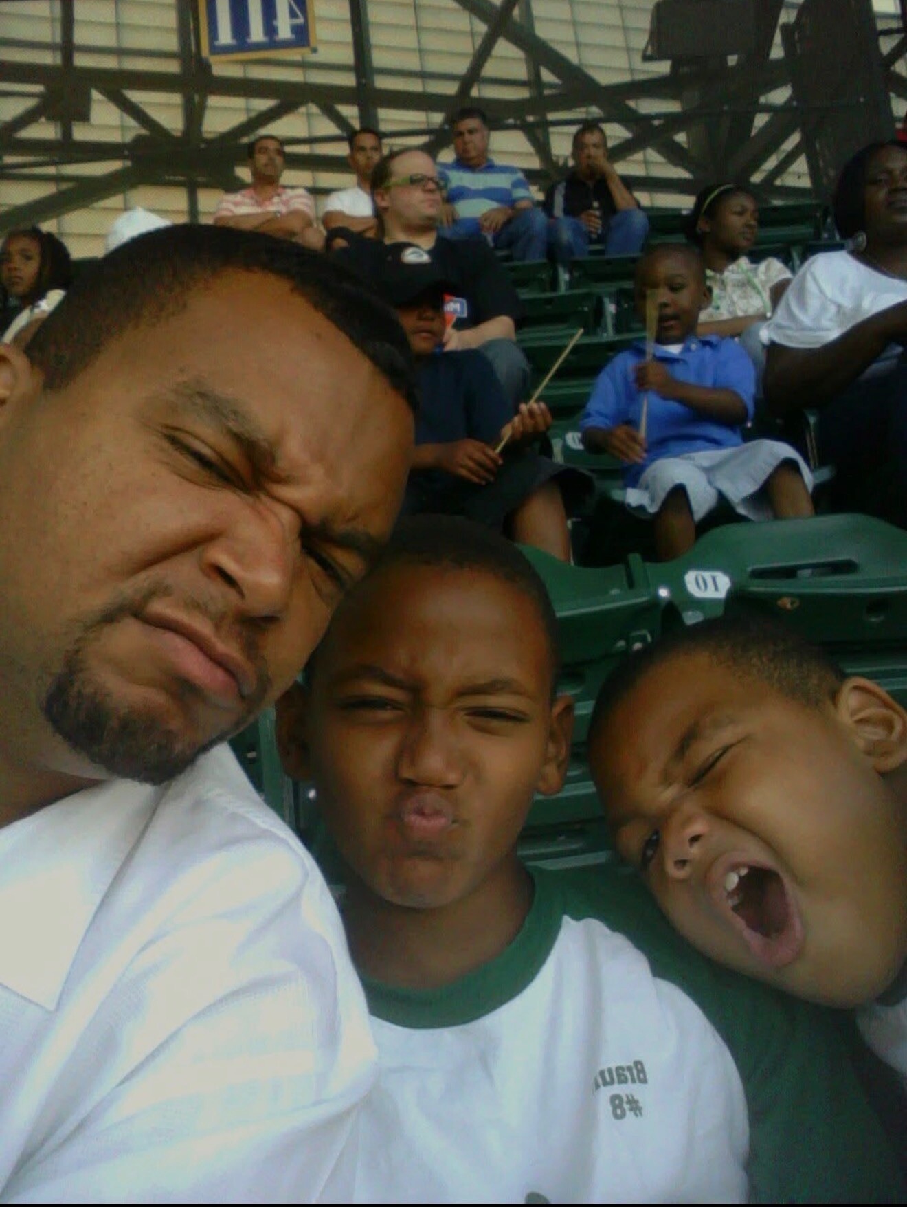 Close-up selfie of a man and two children making funny faces at a baseball stadium, with green bleachers and spectators in the background.