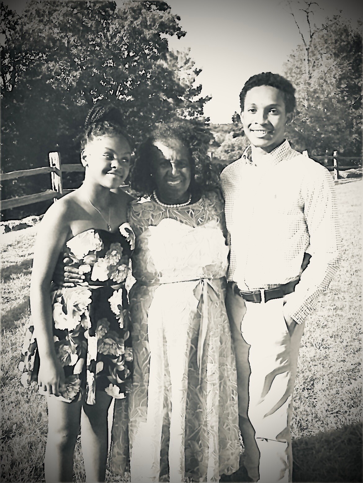 Three people posing outdoors together, two women in dresses and a young man in a button-down shirt, smiling at the camera.