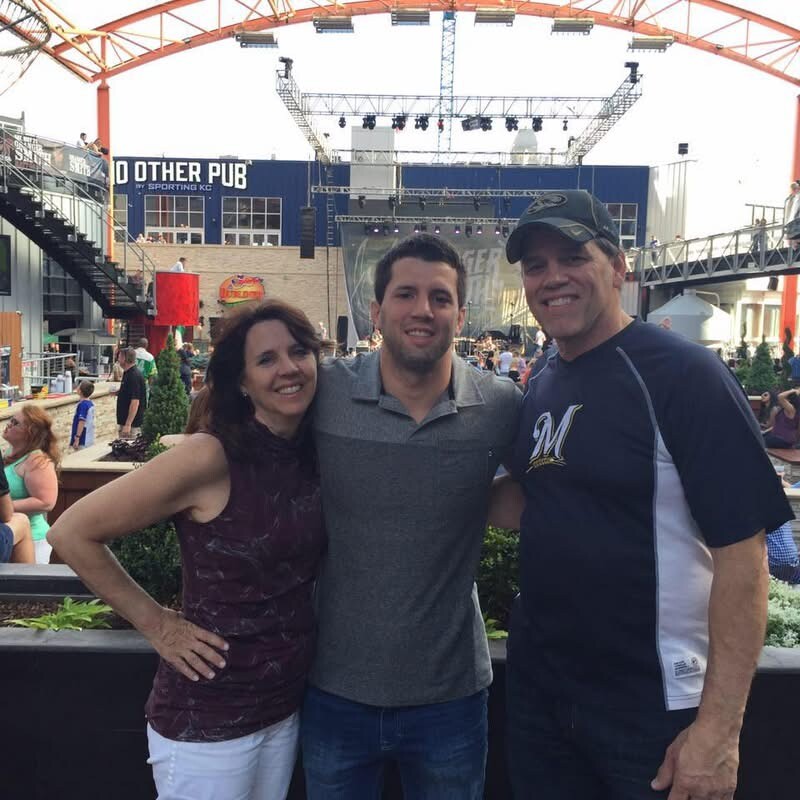 Three adults posing for a casual photo in front of a stage at an outdoor venue, all smiling at the camera.