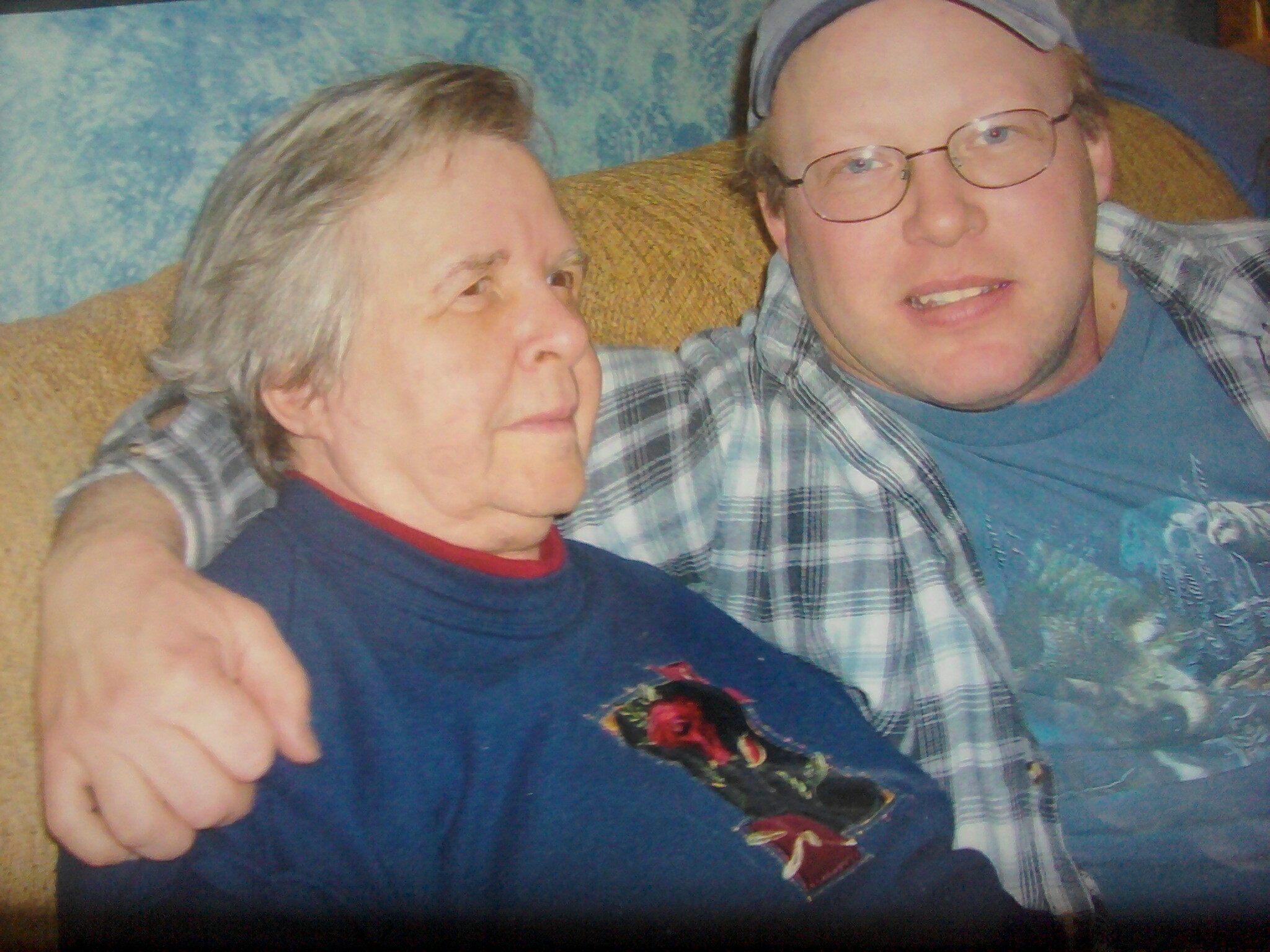 Two people sit close together on a couch, an older woman on the left and a younger man with glasses on the right, smiling softly.