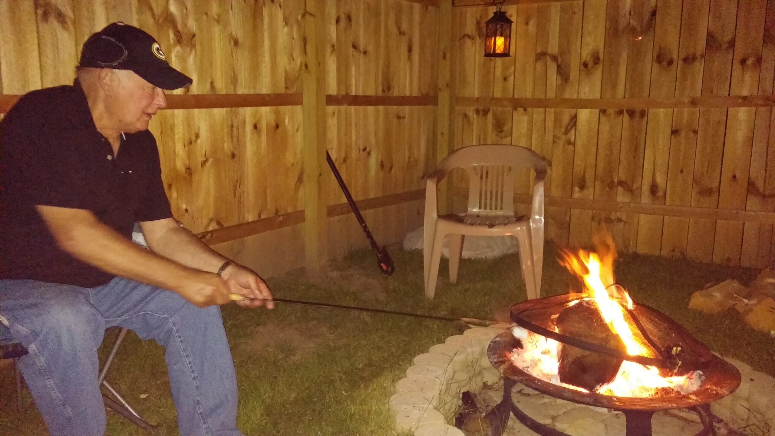 Older man in a dark polo and cap tending a backyard fire in a wooden fenced area at night.