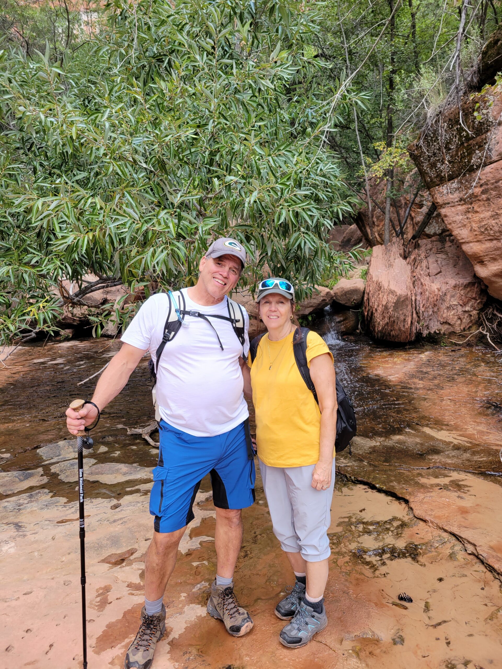Two hikers, a man and a woman, stand side by side in a shallow red-rock stream with dense green foliage behind them, smiling at the camera.