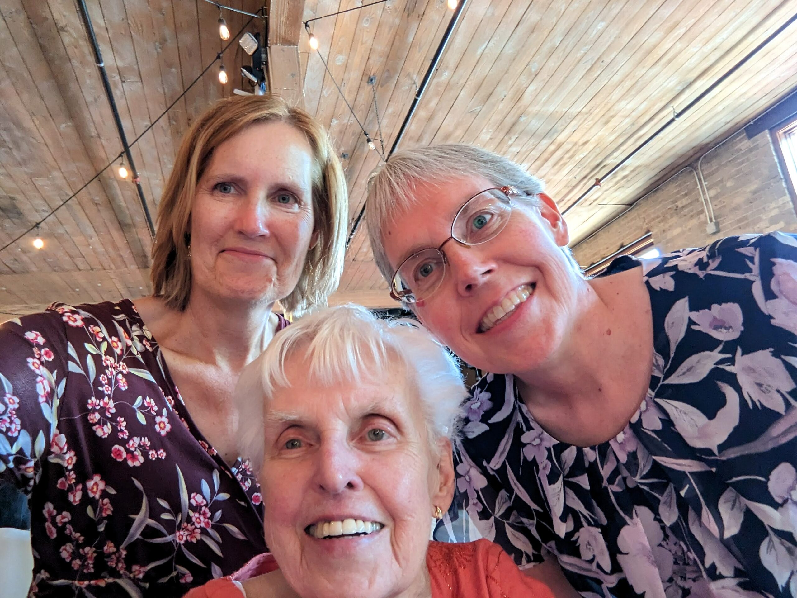 Group of four women posing for a selfie indoors, with a wooden ceiling and string lights in the background.
