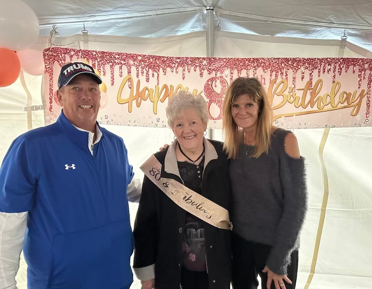 Three people pose for a birthday photo under a pink 'Happy Birthday' banner; the elderly woman in the center wears a sash.