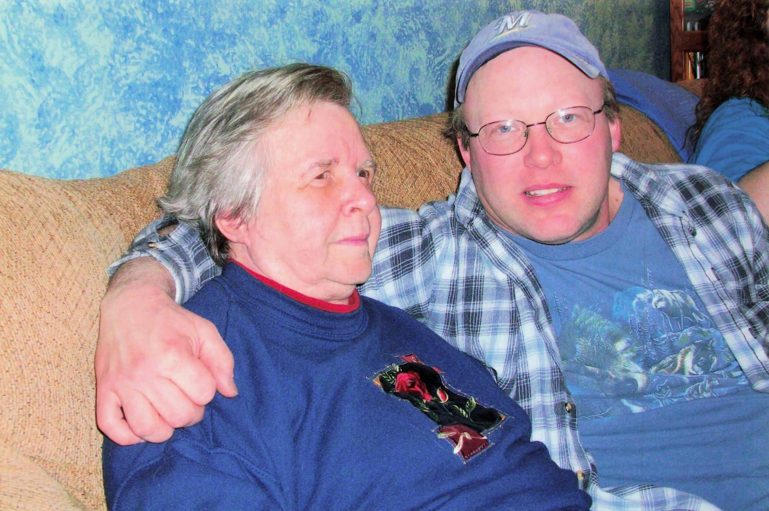 Two men sit close together on a tan couch, one with gray hair and a blue shirt on the left, the other wearing glasses and a light-blue cap on the right, both looking at the camera with smiles.