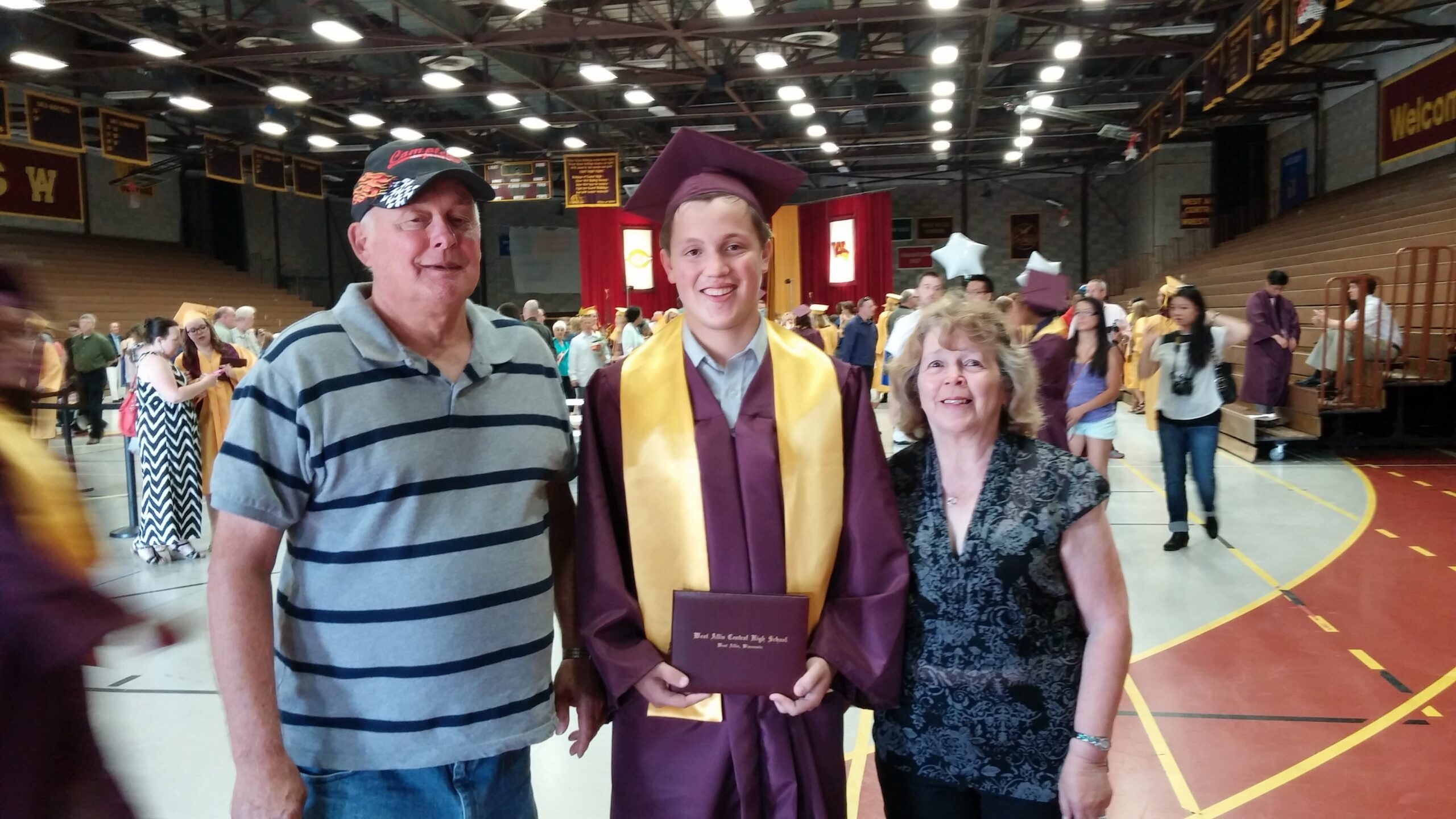 Graduate in maroon gown and cap with yellow stole poses with two relatives in a gymnasium during commencement.