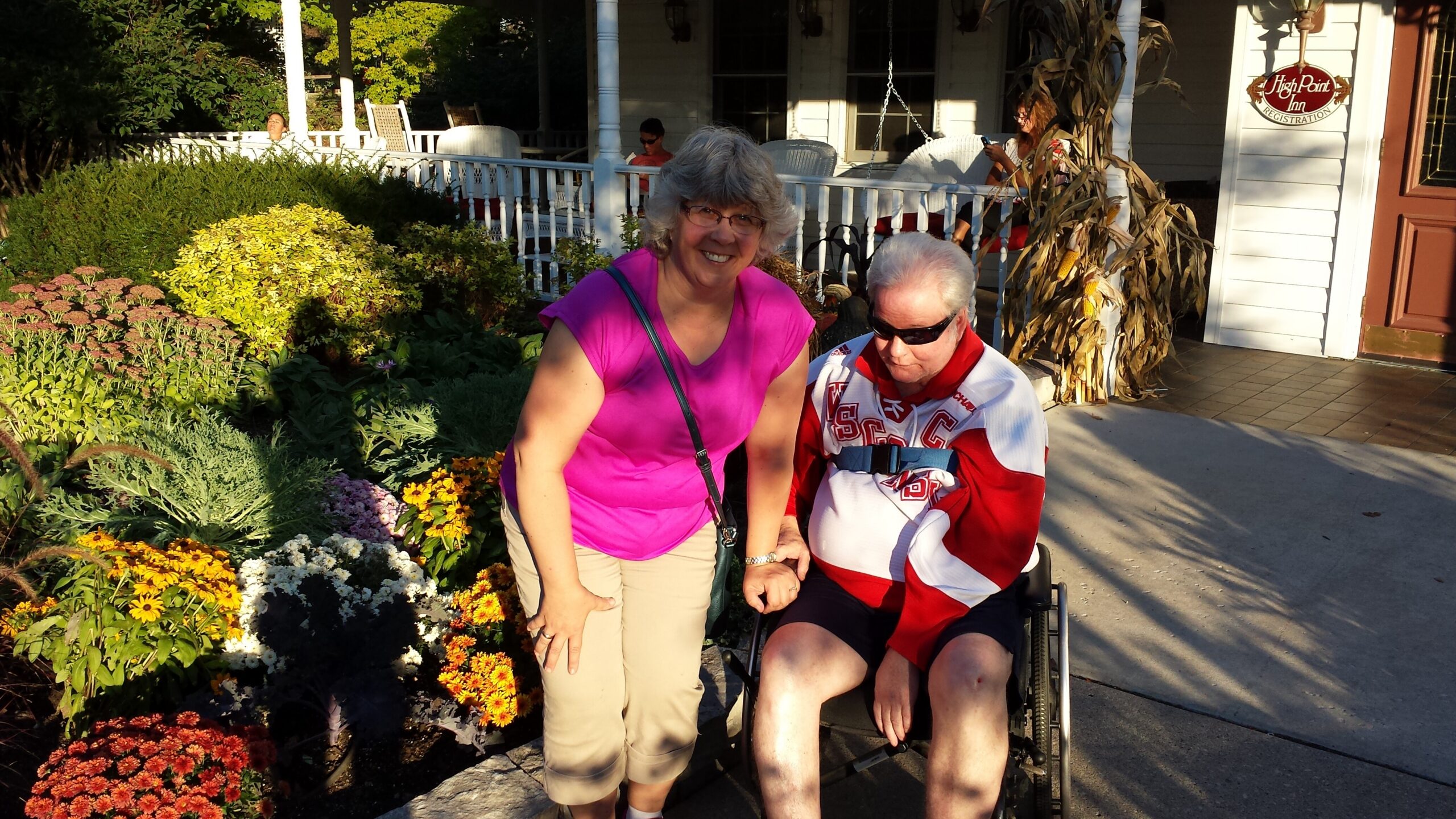 Smiling woman in pink shirt stands beside a man in a wheelchair among colorful garden flowers in front of a porch.
