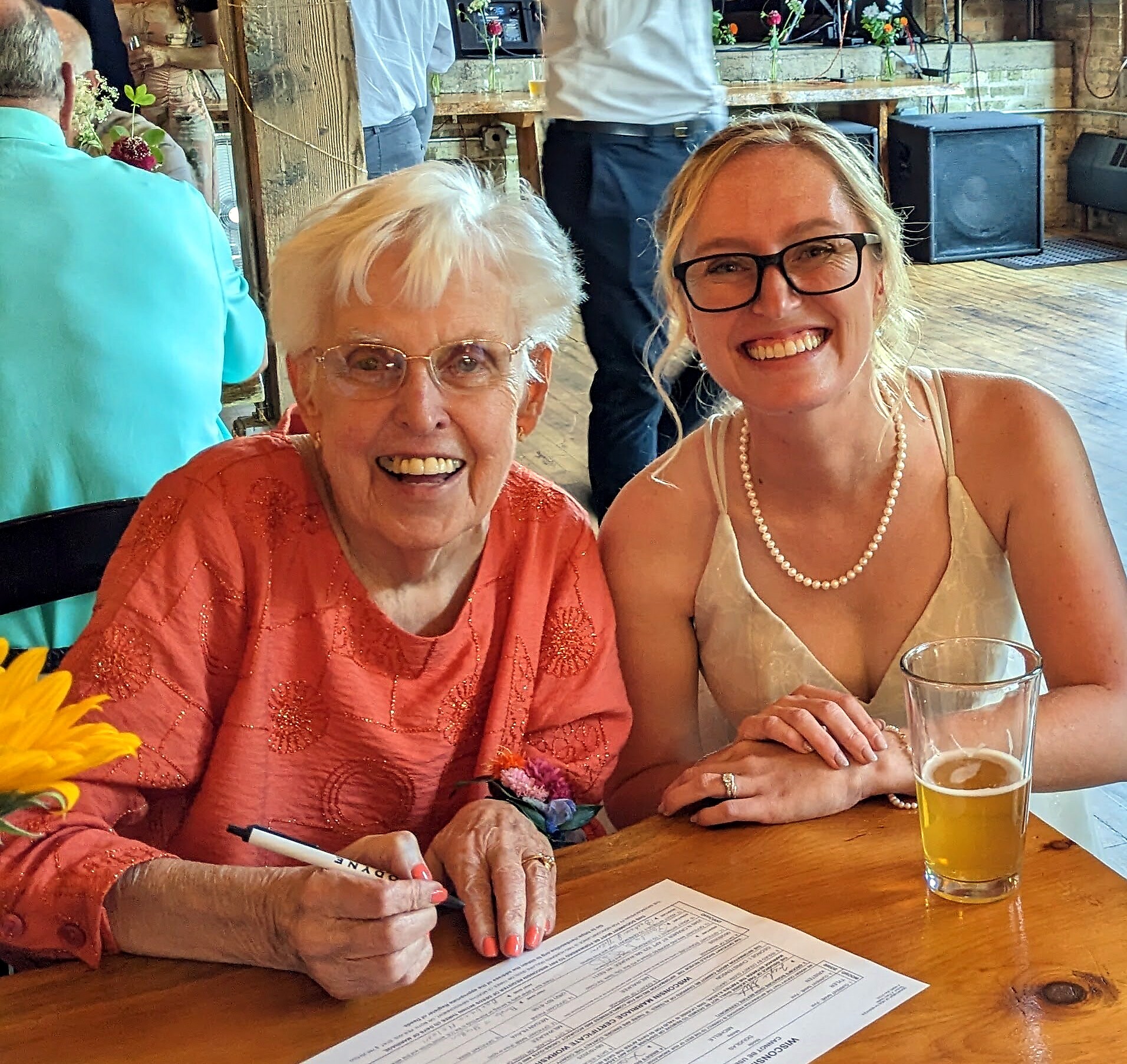 Two smiling women sit at a wooden table; older woman signs a document while the younger woman grins beside her, with a beer glass in view and a menu on the table.