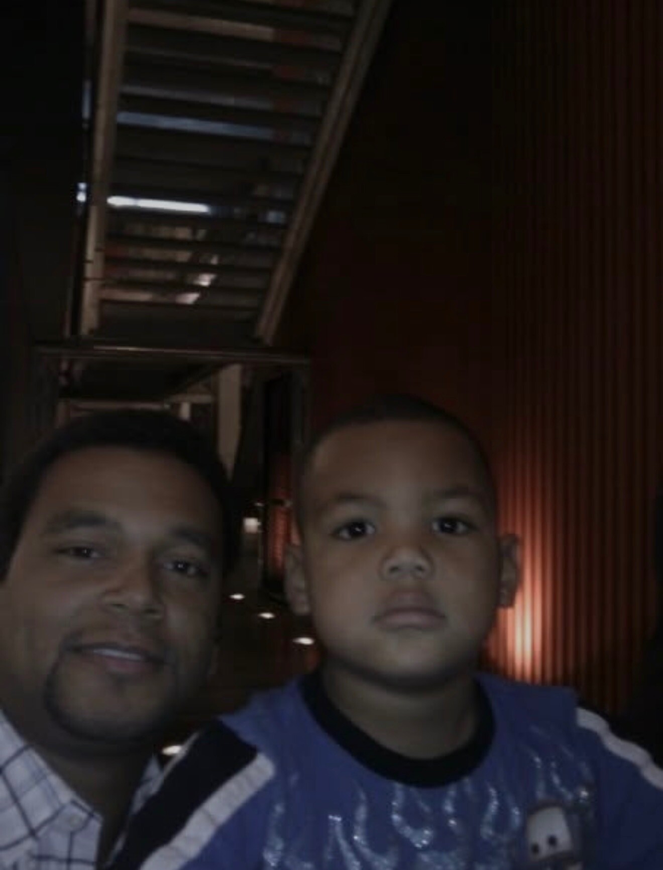 Man and young boy taking a selfie in a dimly lit indoor hallway/similar setting.