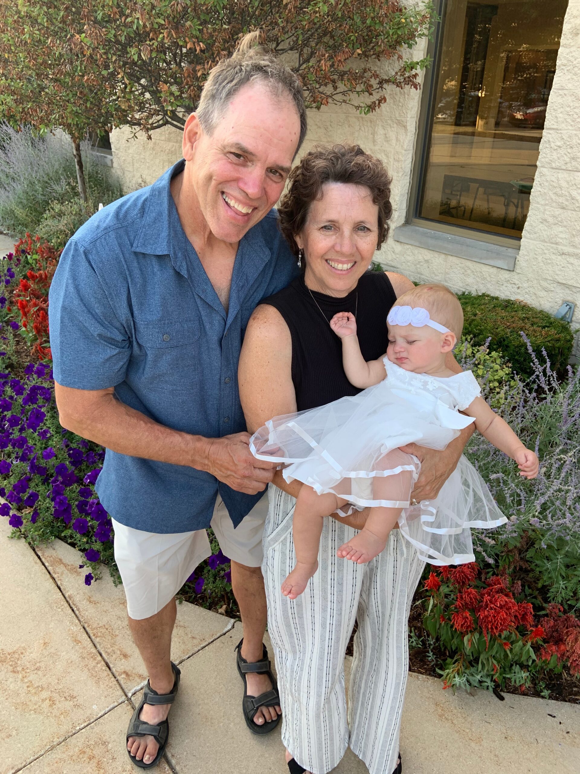 Smiling couple holding a baby girl in a white dress outdoors beside a flowerbed of purple and red flowers.