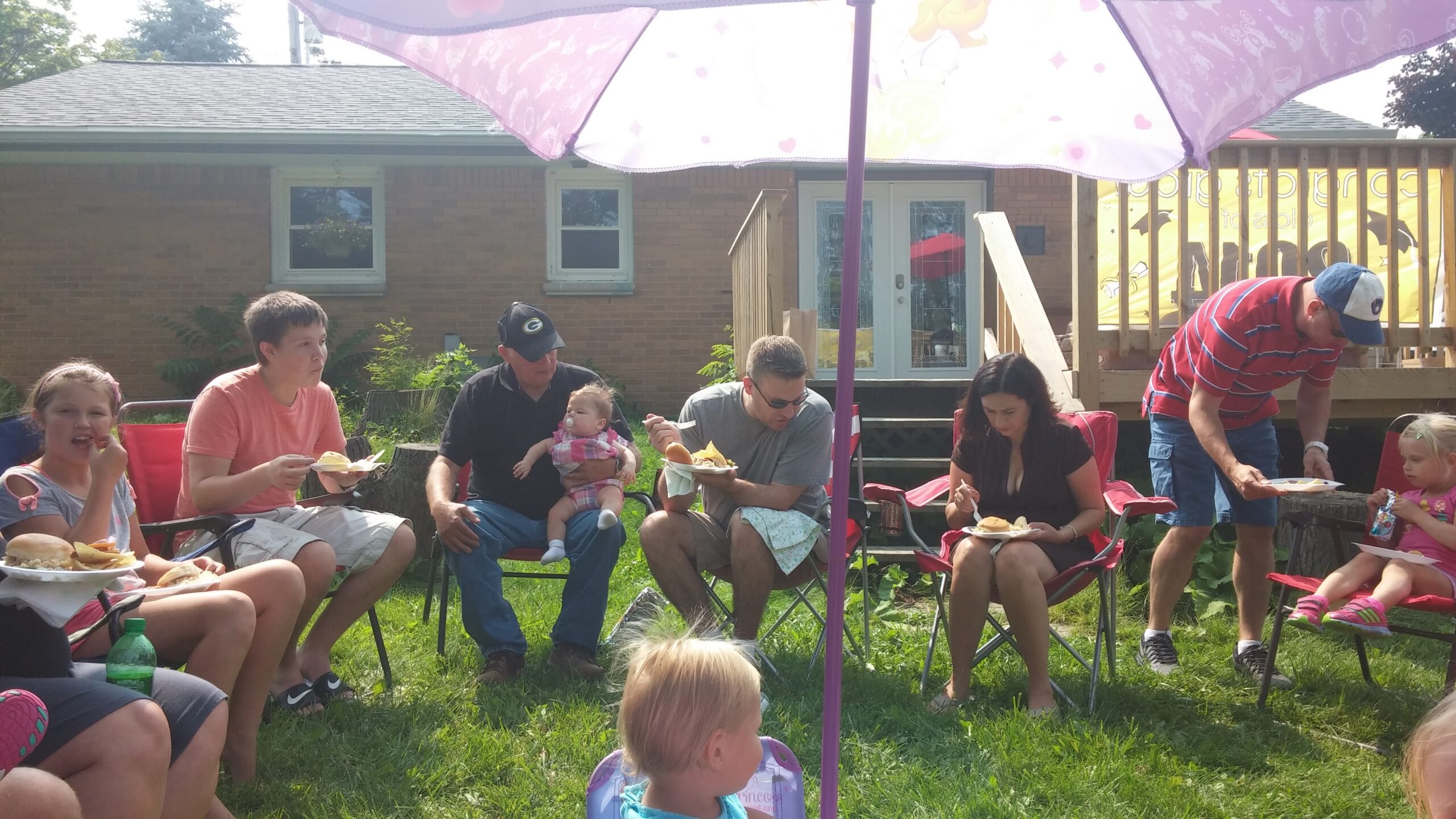 Group of family and friends having a backyard cookout with burgers under a pink patio umbrella.