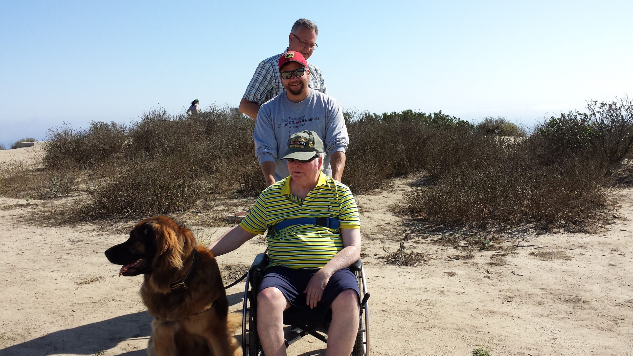 Man in a wheelchair on a sunny desert trail with a dog in the foreground and a companion standing behind him.