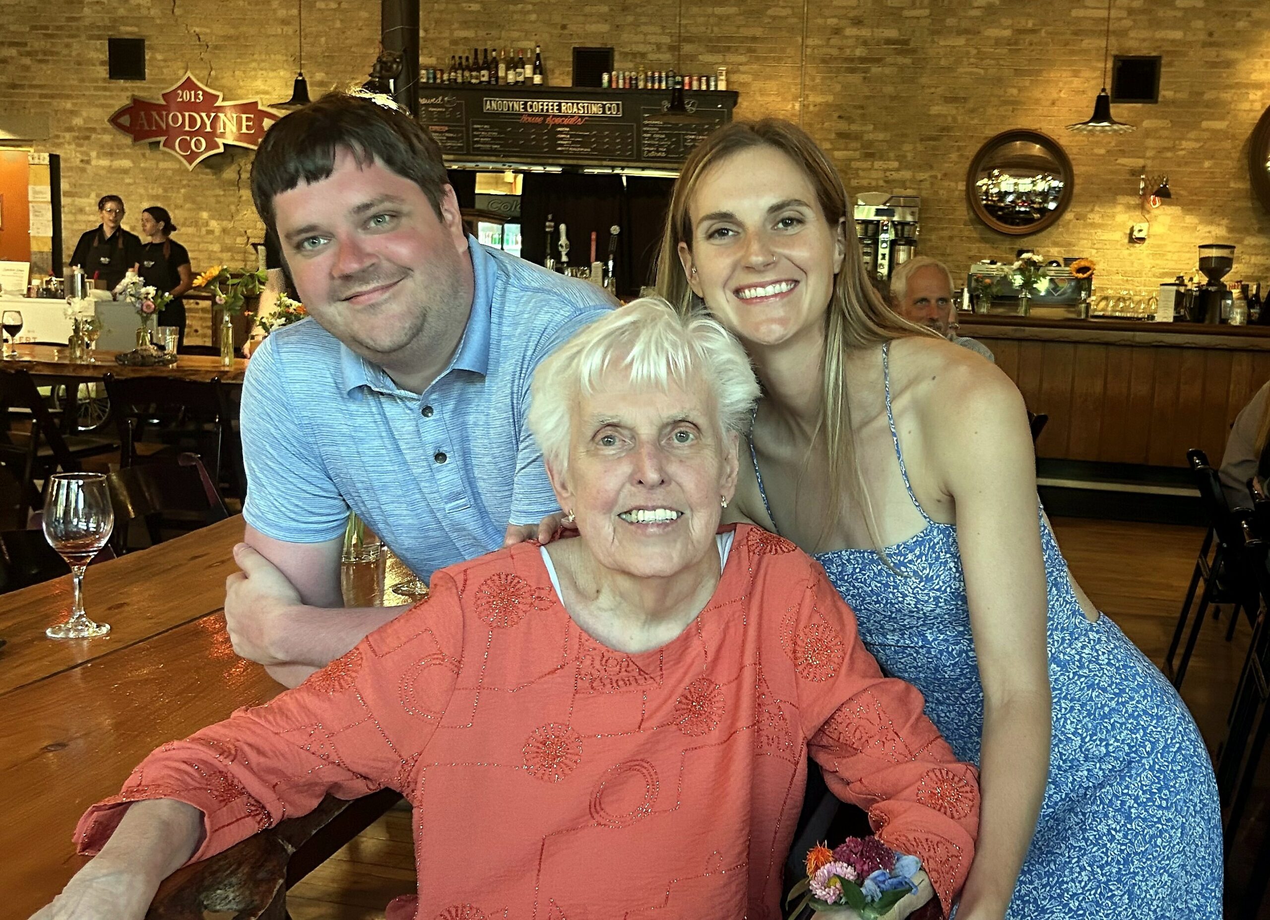 Three people pose together at a wooden restaurant table; an elderly woman in an orange top sits center, flanked by a man in a blue shirt and a smiling woman in a blue patterned dress.