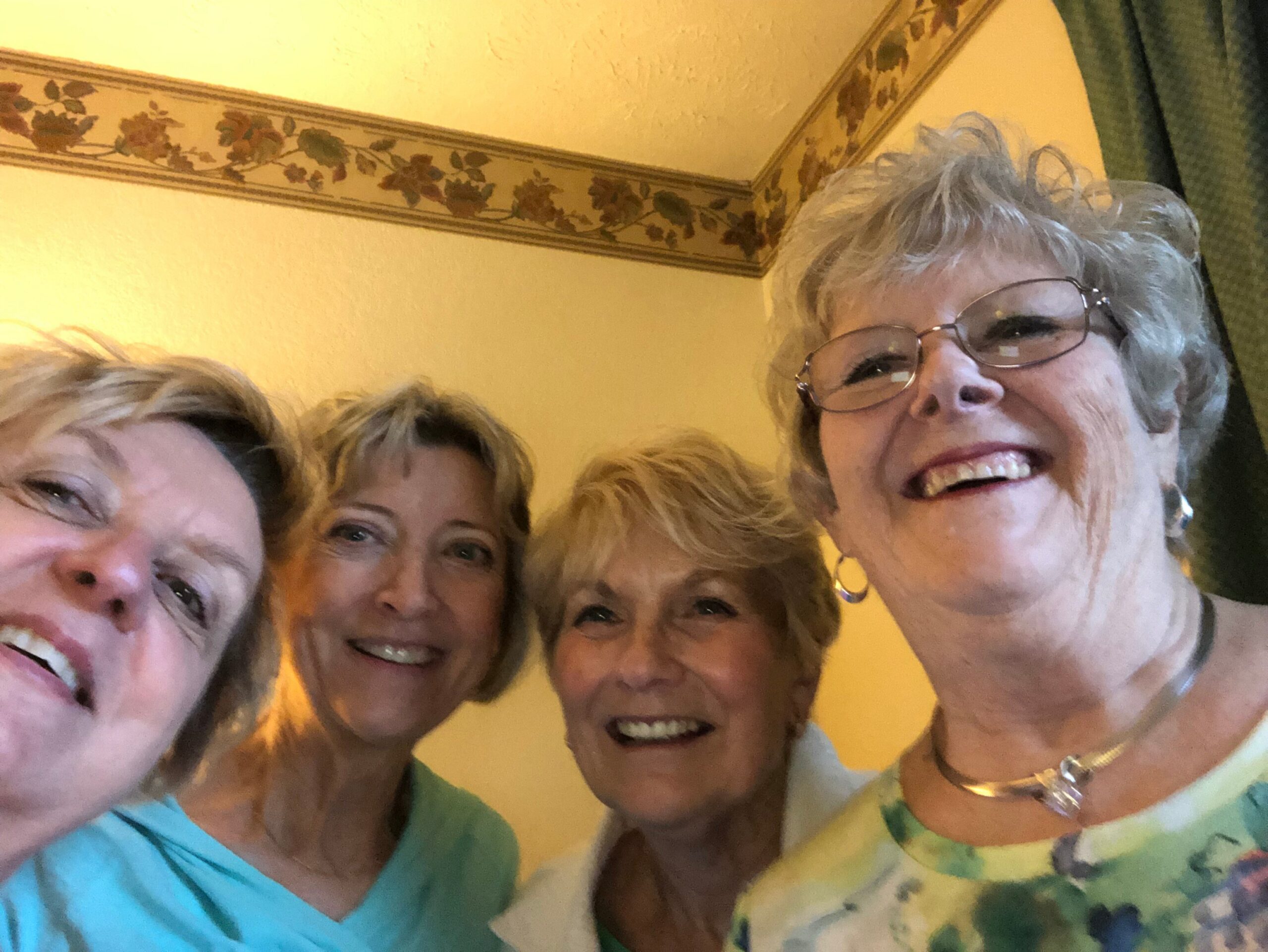 Five smiling older women posing for a close-up group selfie indoors, faces filling the frame.