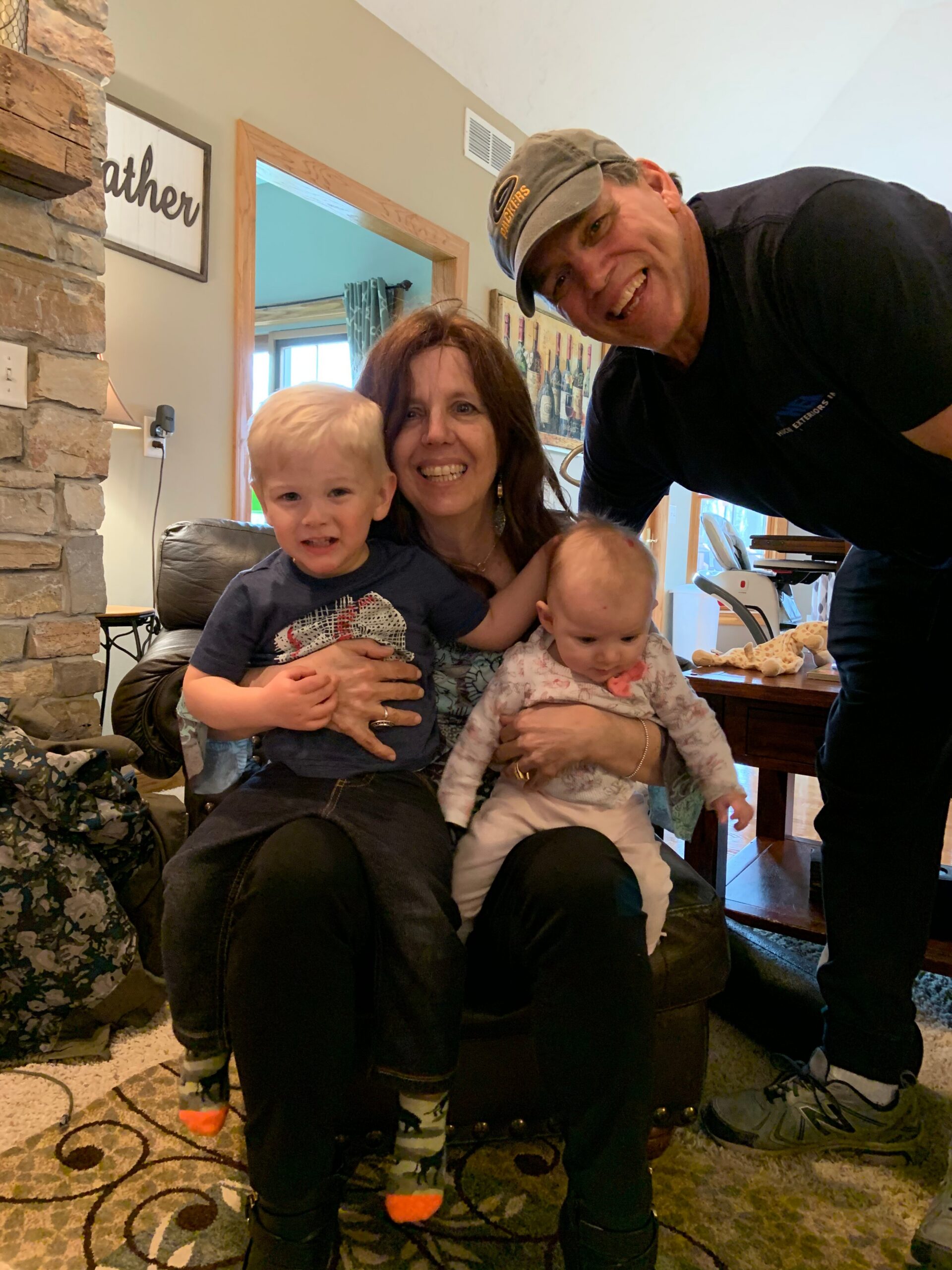 Mother sitting on a sofa holding two young children, all smiling at the camera indoors with a man leaning in from the right.