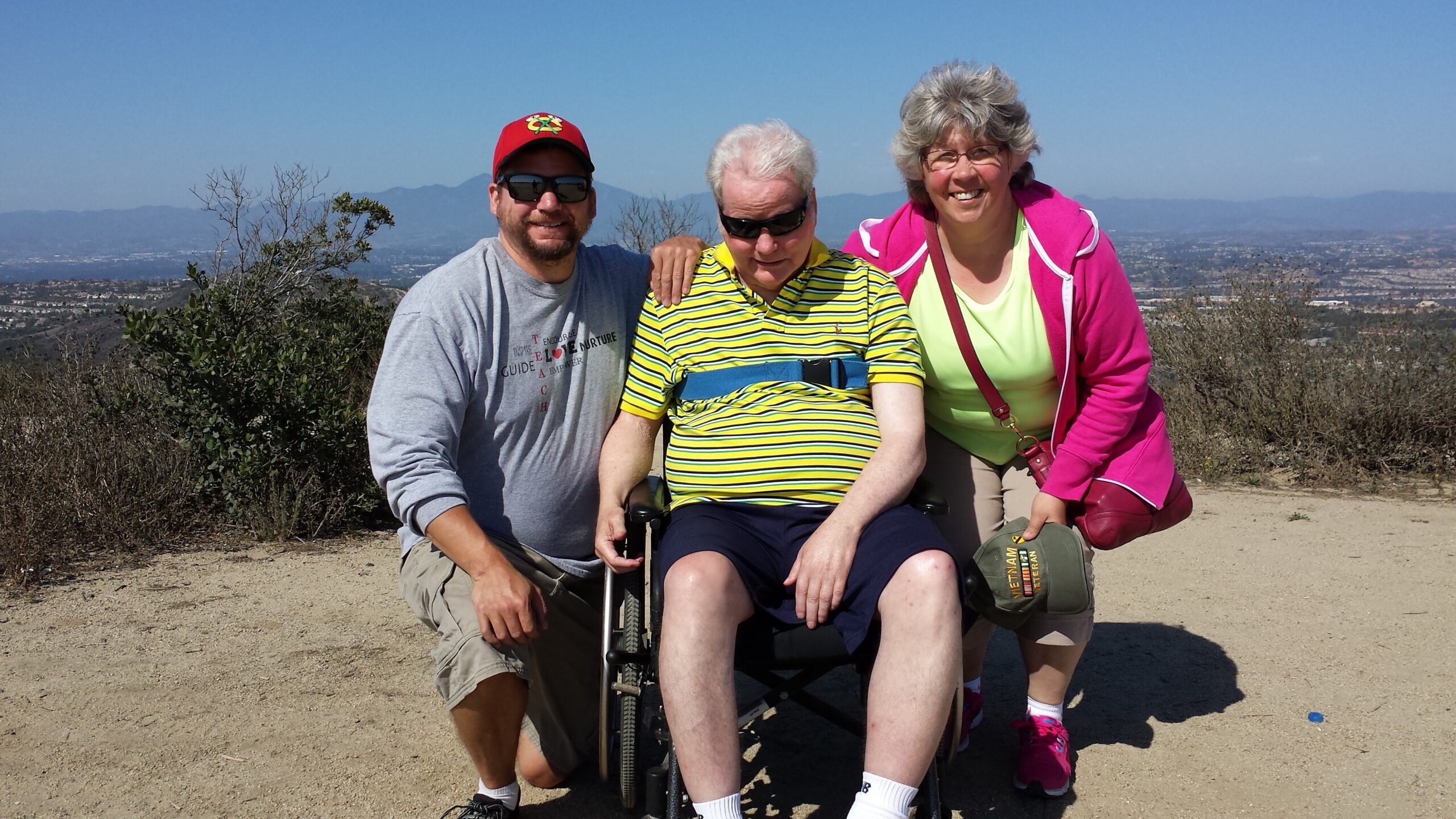 Three people outdoors on a hilltop: a man in a gray shirt beside a woman in a wheelchair, with a smiling woman in a pink jacket beside them; scrubby landscape and distant city.