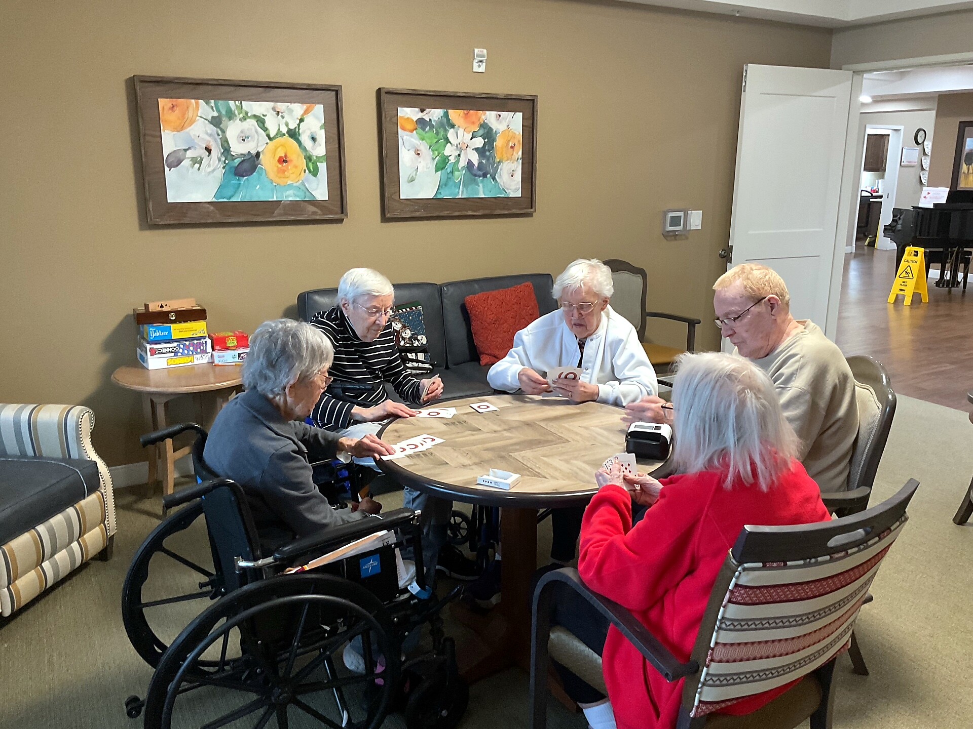 Group of elderly residents around a round table playing cards in a lounge, some in wheelchairs, with paintings on the wall and a casual, cozy atmosphere.