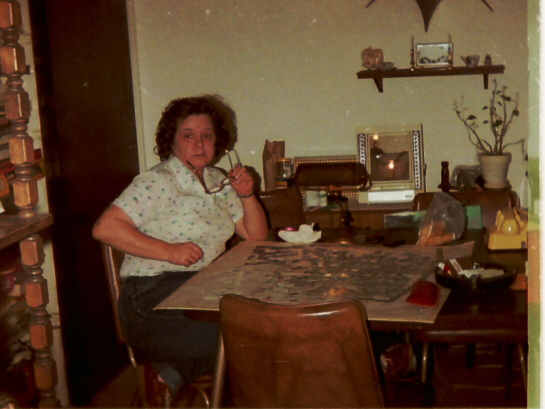 Older woman sits at a dining table with a jigsaw puzzle spread out, holding glasses near her face in a retro-style room.