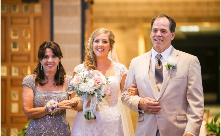 Bride in a white lace gown walks with her parents; bouquet in hand, smiles as they pose together at the wedding.