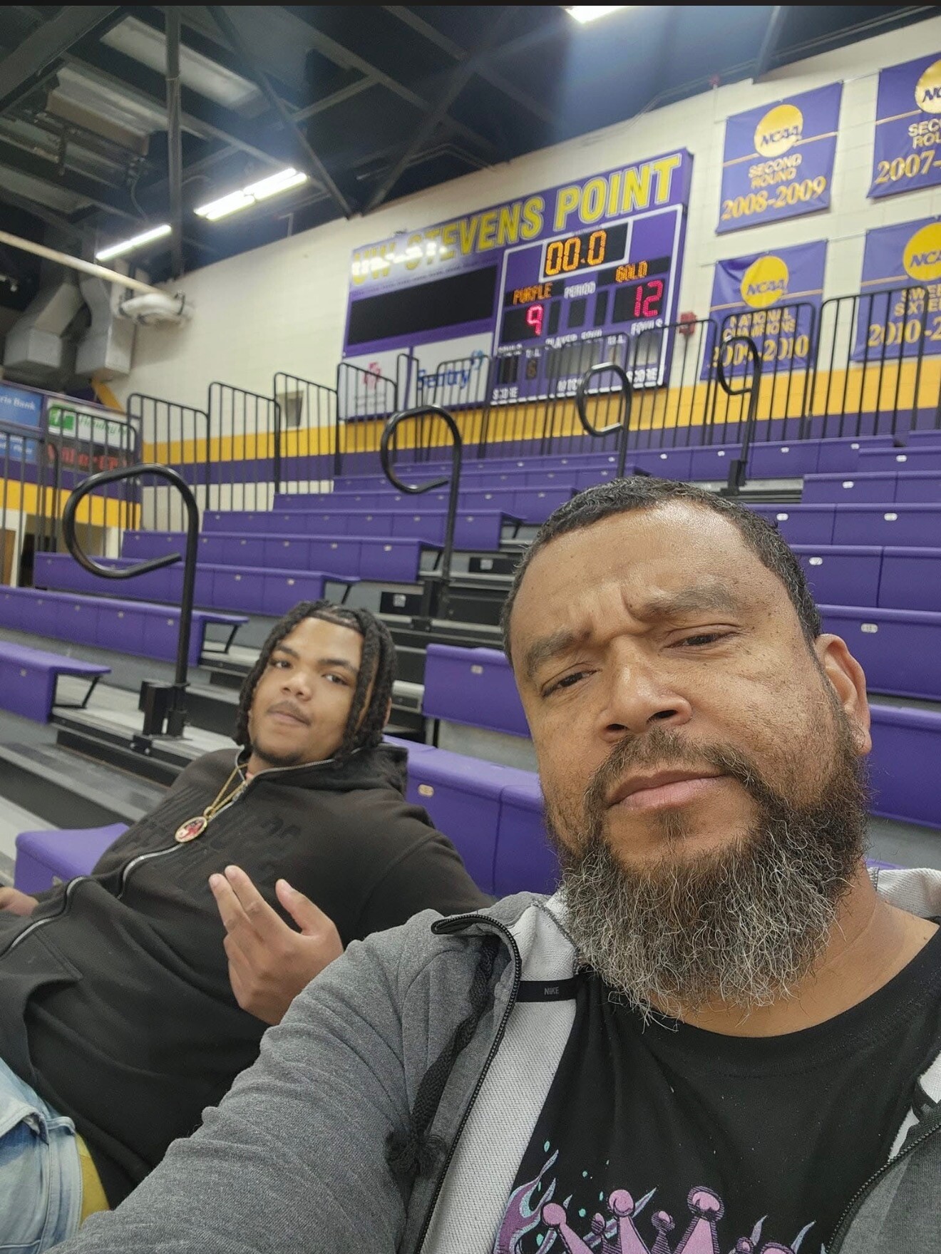 Selfie in a gym: bearded man in front and a man with braided hair seated in purple bleachers, scoreboard and banners visible in the background.