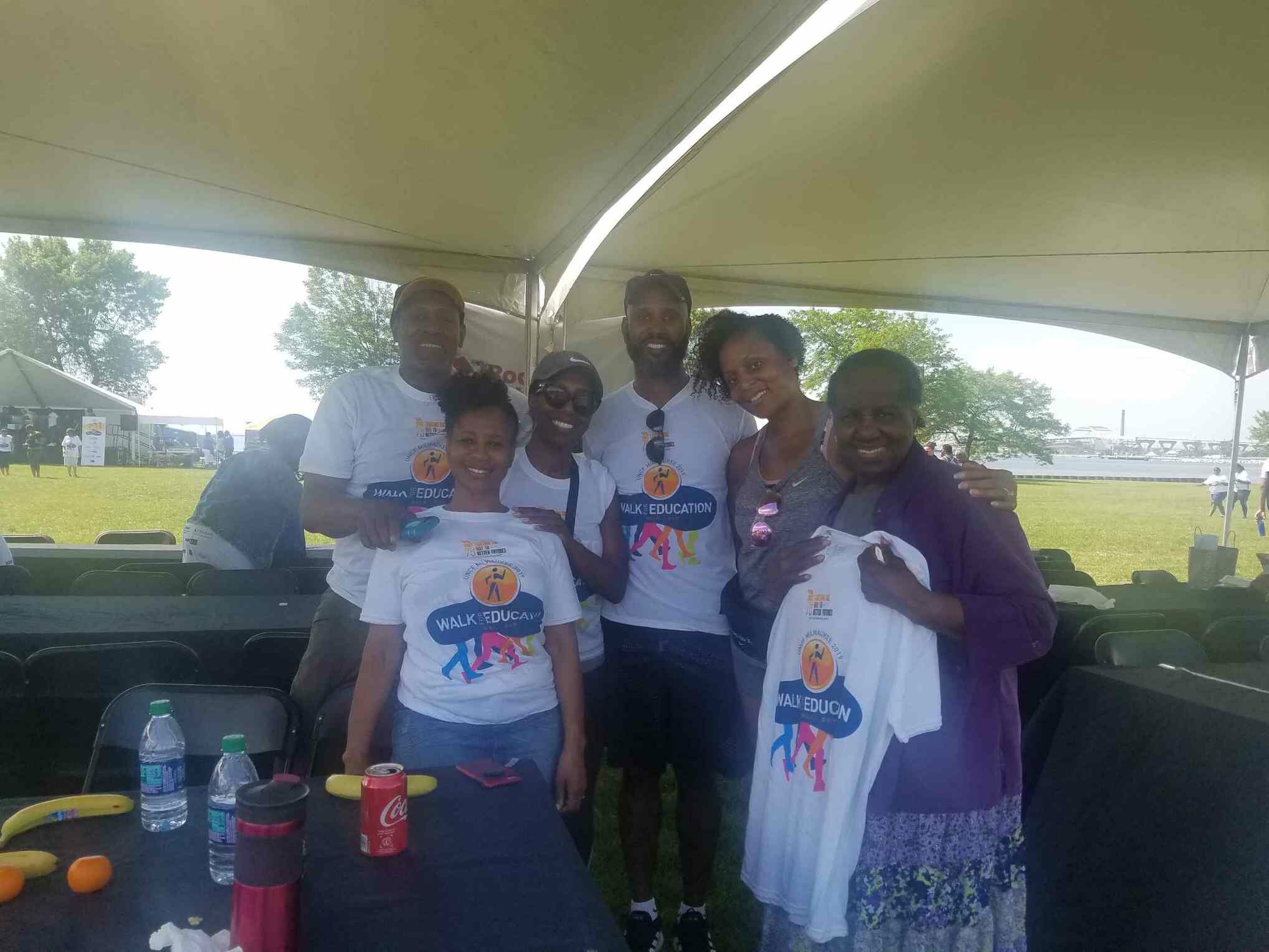 Group of smiling volunteers under a large white canopy at an outdoor event, wearing Walk Education shirts.