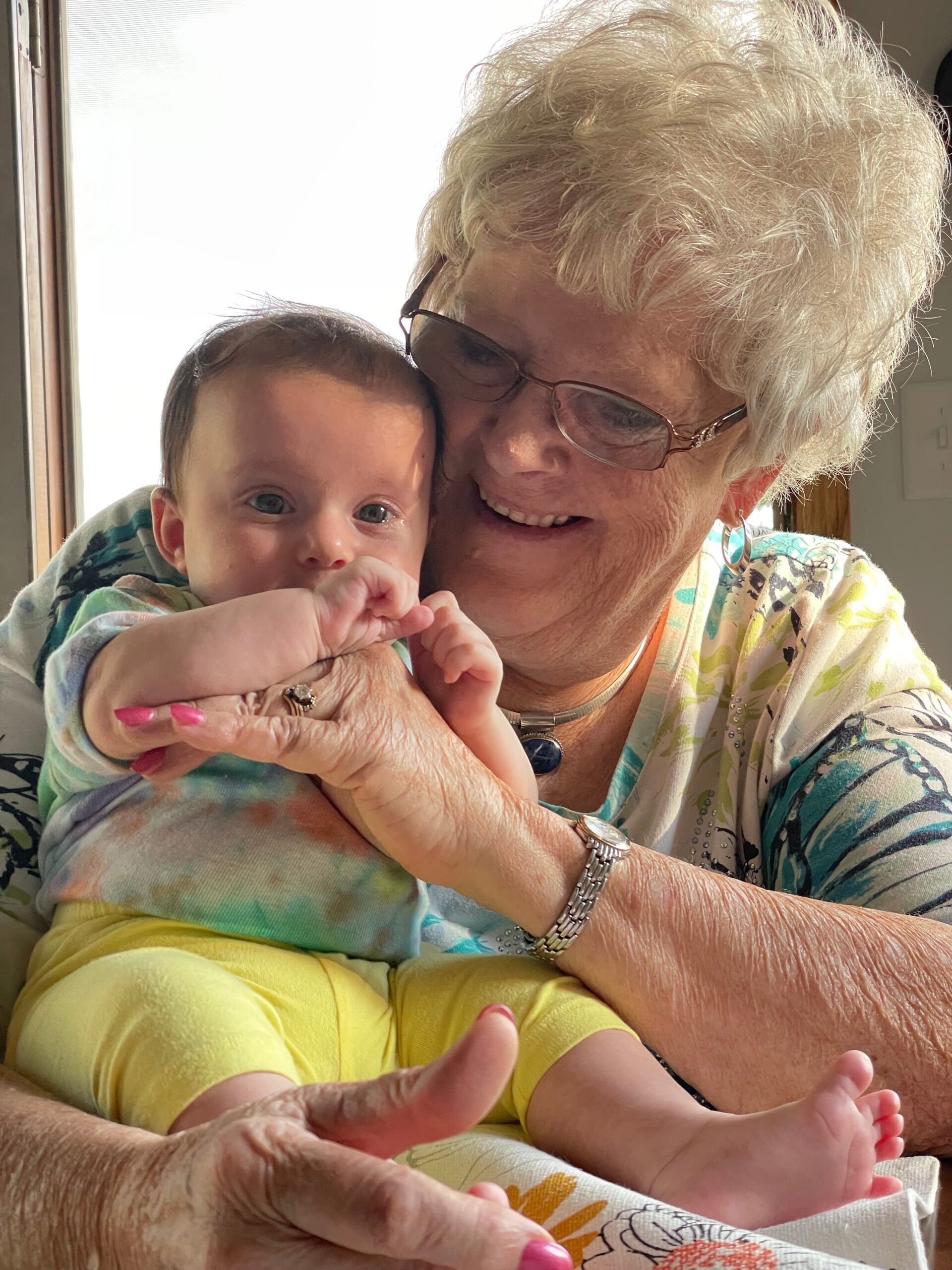 Grandmother with short gray hair and glasses smiling while holding a baby in bright clothing near a windowulai.”