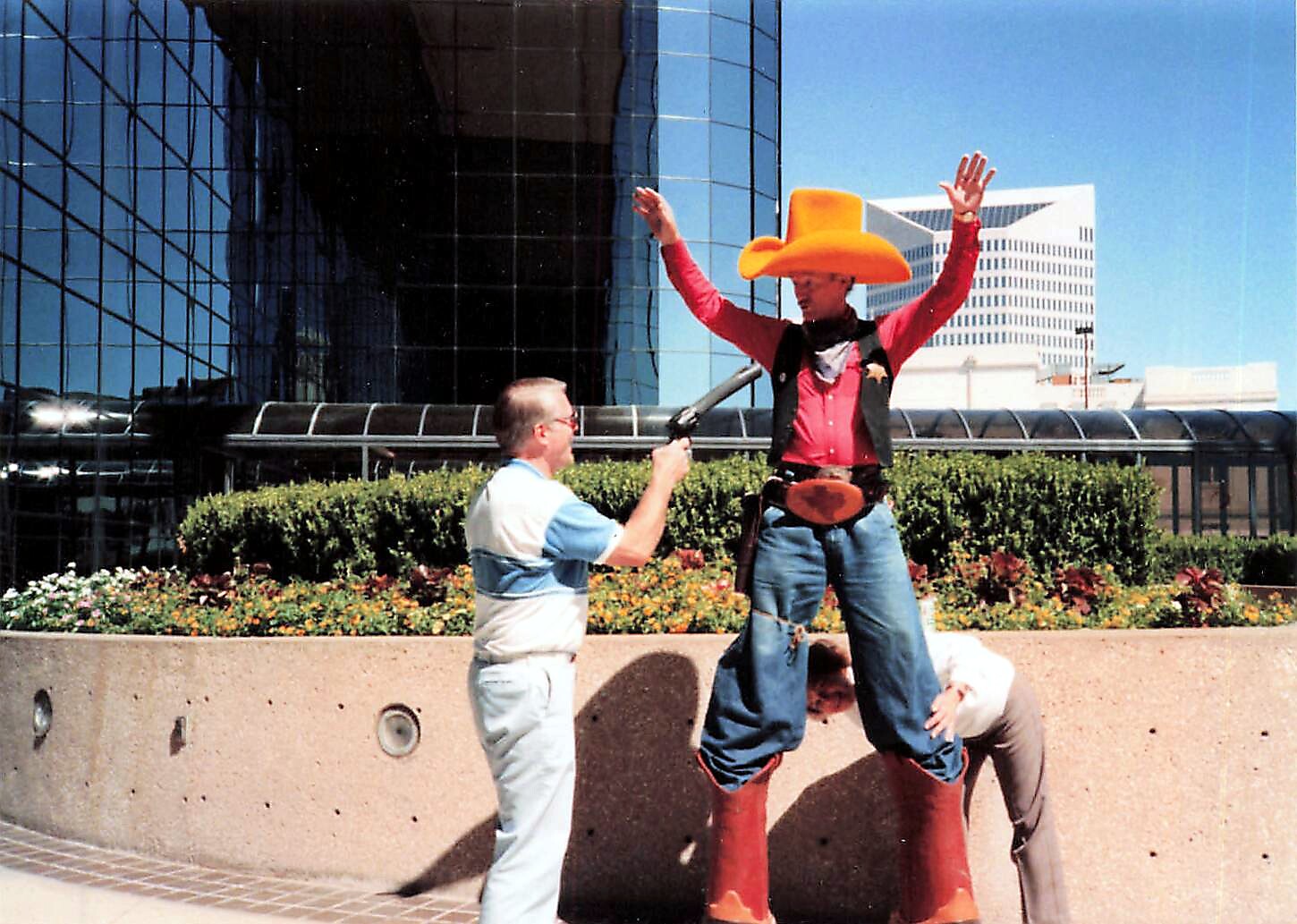Stilt-walker in a cowboy outfit with a huge orange hat raises his arms as a man with a microphone talks to him in a city plaza with glass buildings in the background.