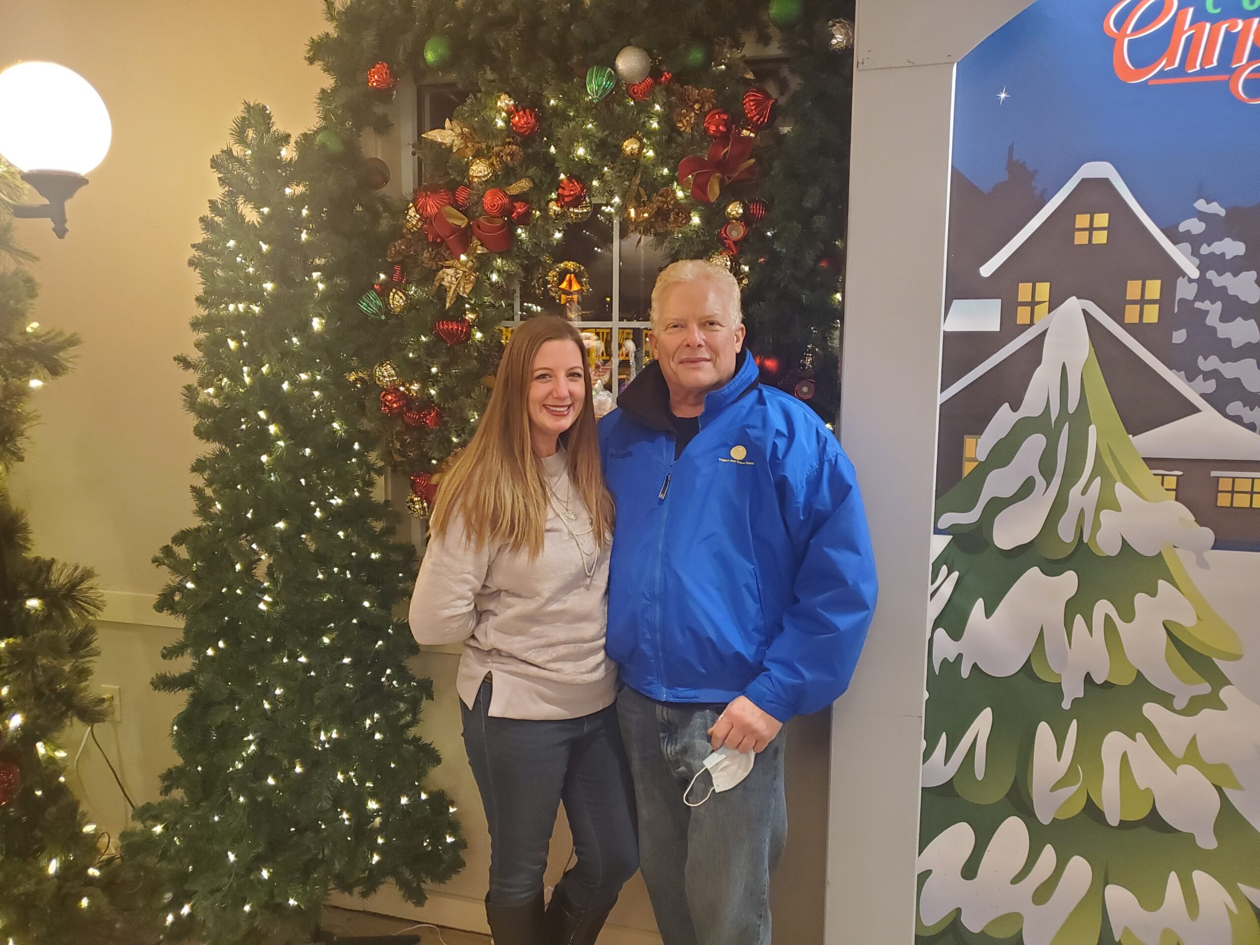 Two people smile for a photo beside a lit Christmas tree with red and gold ornaments indoors.