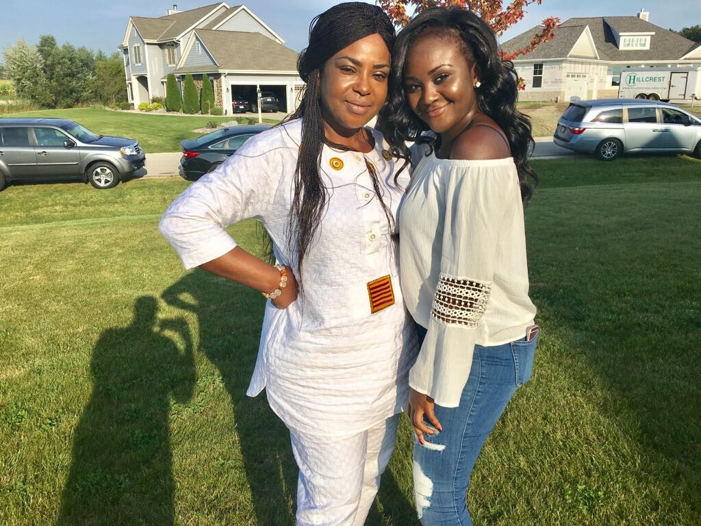 Two women posing together on a sunny lawn in a suburban neighborhood; one wears a white traditional outfit, the other a white off‑the‑shoulder top and jeans.