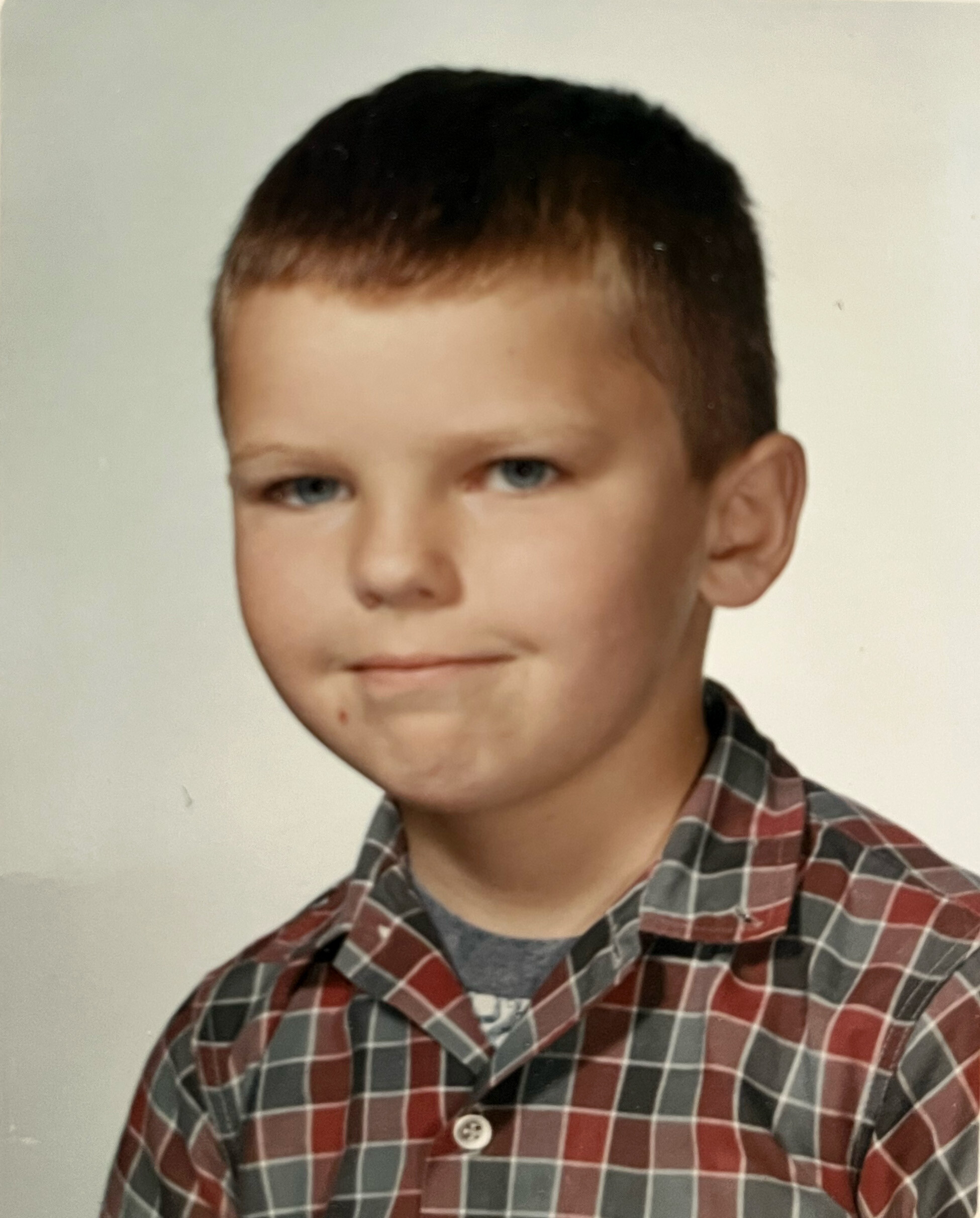 Partial portrait of a young man wearing a red and gray plaid shirt, facing slightly left, with neutral expression.