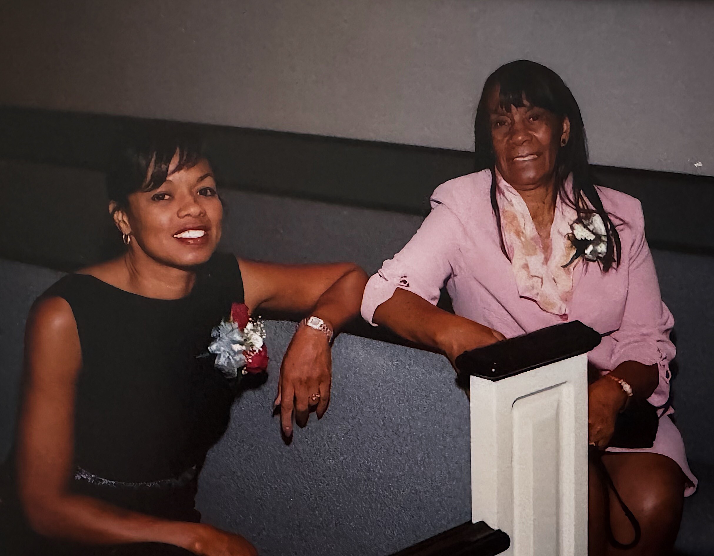 Two women posing on a stair-step ledge indoors, smiling at the camera; one in a black dress with a corsage and the other in a pink blazer.