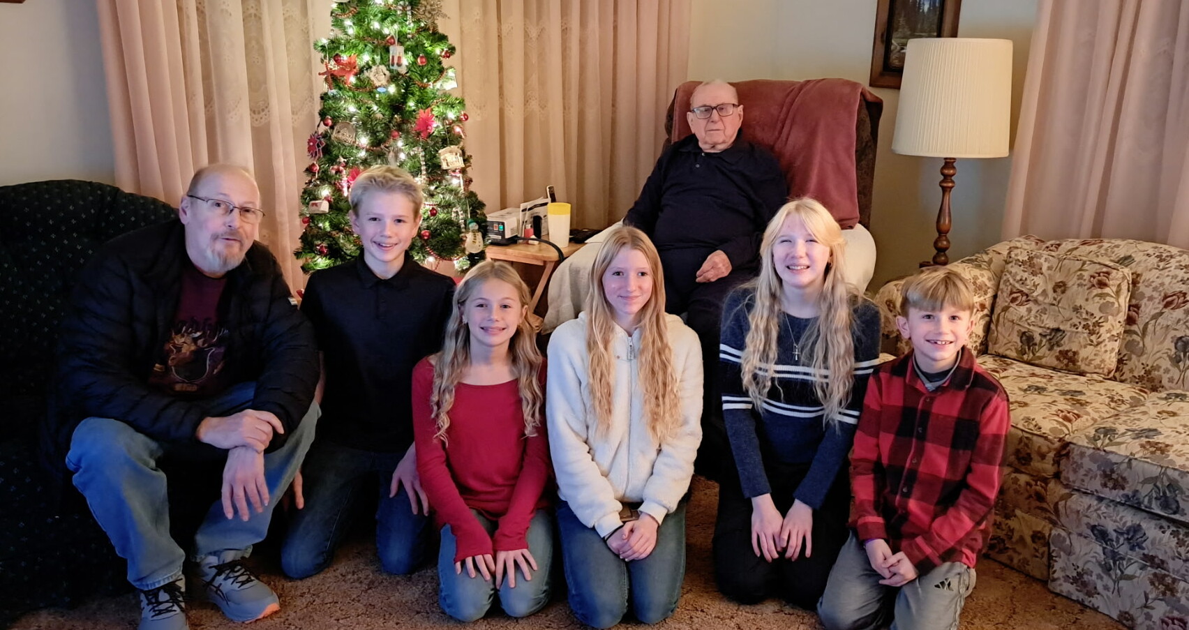 Six family members pose for a Christmas photo in a living room beside a decorated tree.