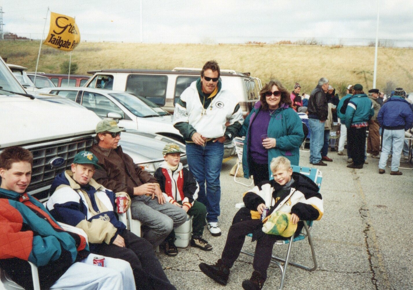 Group of fans tailgating in a parking lot with cars and a yellow Tailgate flag, adults and kids sitting and standing, holding drinks.